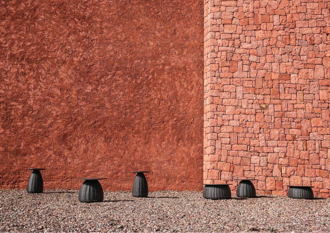 Dedon Porcini Beistelltische, Ø61cm, aus Outdoor-Geflecht vor roter Wand und Steinen. Gartenmöbel für Terrasse.