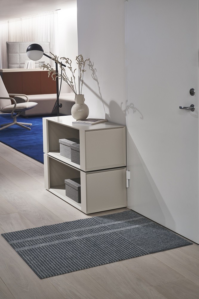 Løype Heymat Fußmatte, grau gestreift, vor einer weißen Tür und einem hellen Regal.