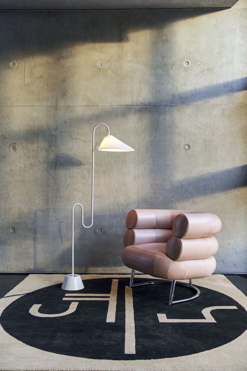 Weiße Roattino Stehleuchte von ClassiCon neben Sessel auf modernem Teppich vor Betonwand.