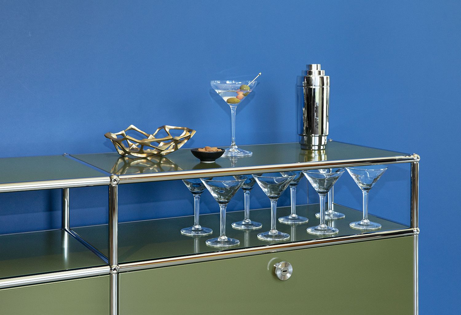 USM Haller Sideboard in Grün mit Cocktailgläsern und Barzubehör auf Glasablage vor blauer Wand.