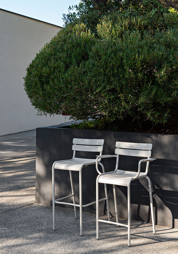 Zwei graue Luxembourg Barhocker von Fermob im Freien vor einer Hecke und Mauer.