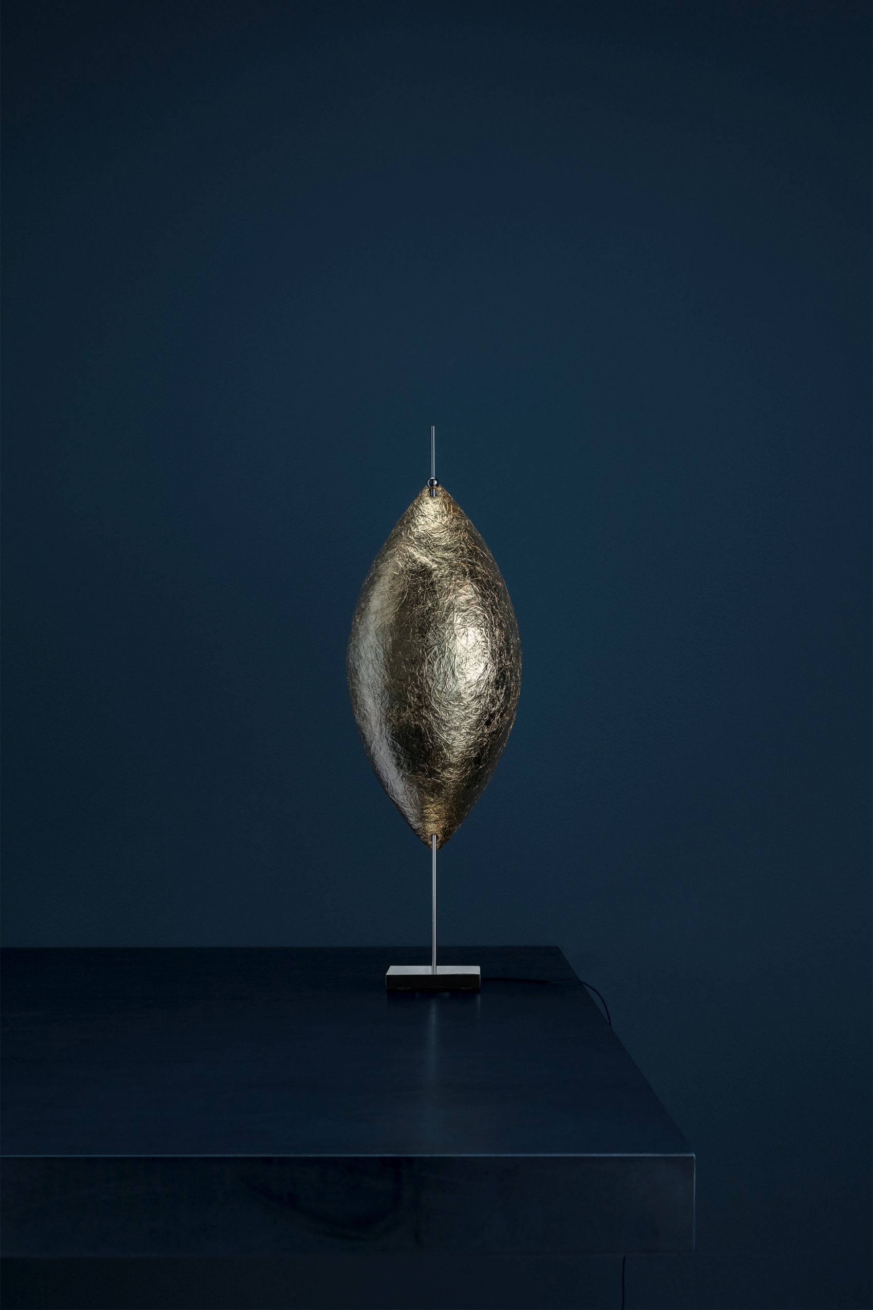 Malagolina Tischleuchte von Catellani & Smith in Gold: Moderne Lampe mit Blattgold-Effekt auf dunklem Tisch.