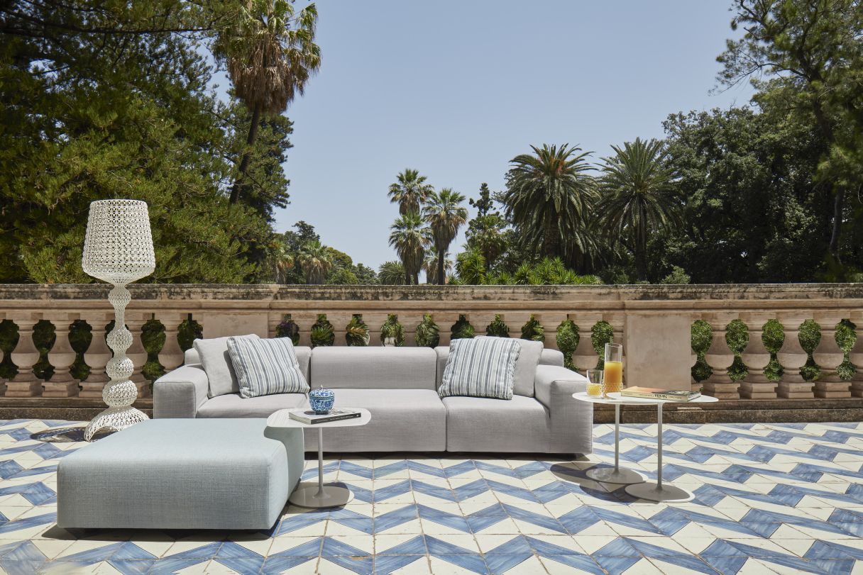 Graues modulares Outdoor-Sofa mit Kissen, Hocker und Beistelltischen auf einer Terrasse mit Palmen im Hintergrund.