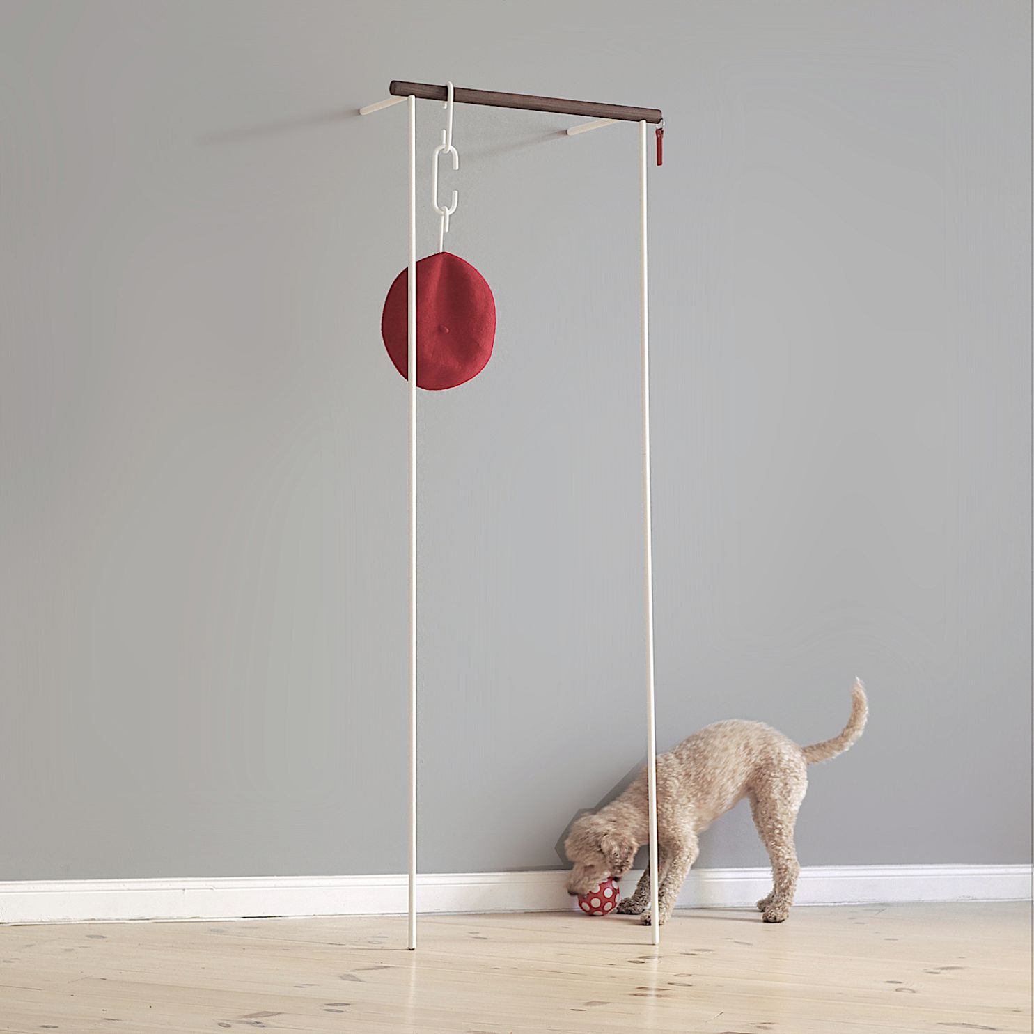 Weiße Anlehngarderobe mit Nussbaumstange, rotem Hut und Hund mit Ball im Bild.