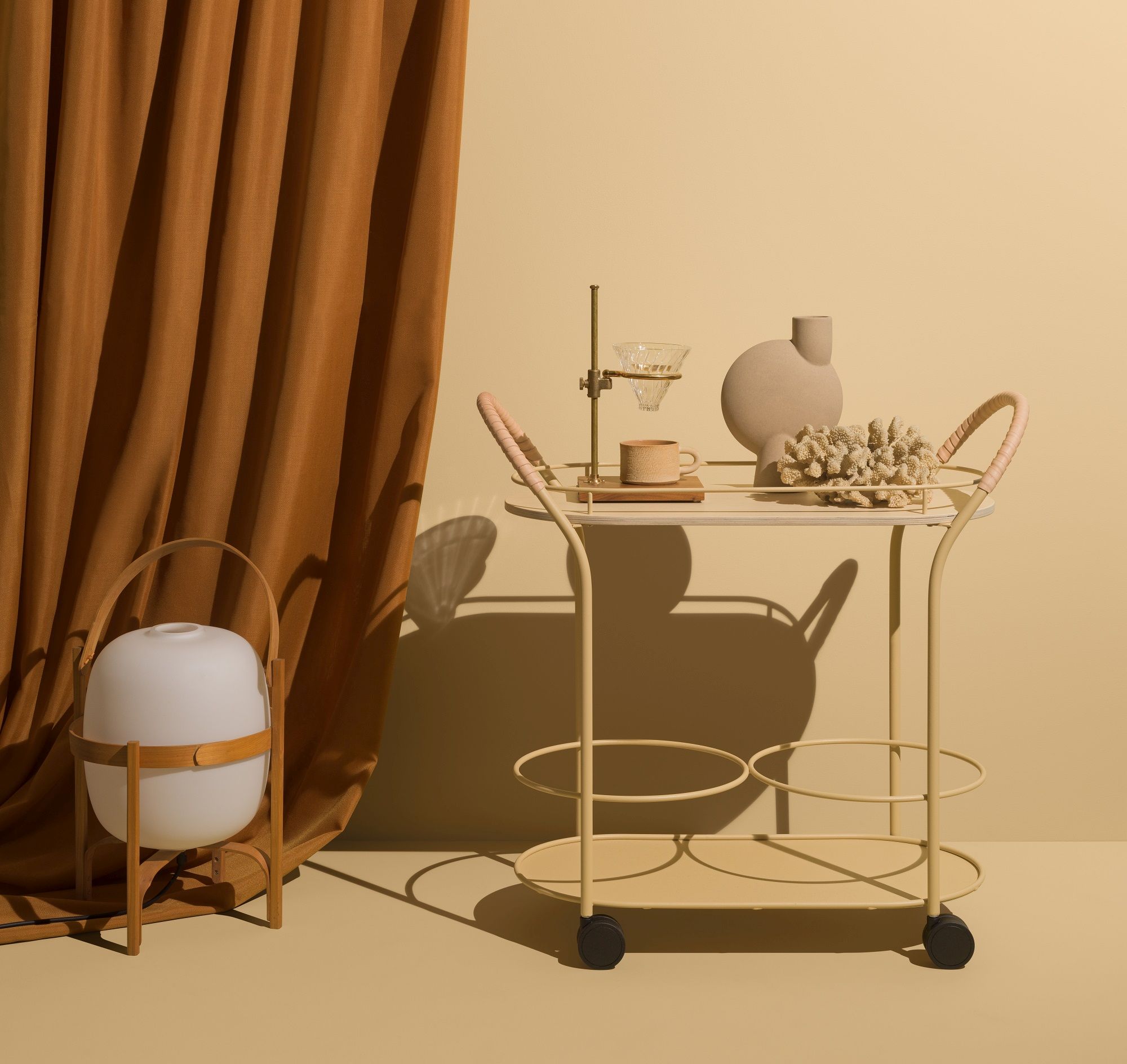 Beiger Bistro Servierwagen von Mizetto mit Deko-Objekten und Lampe vor braunem Vorhang.