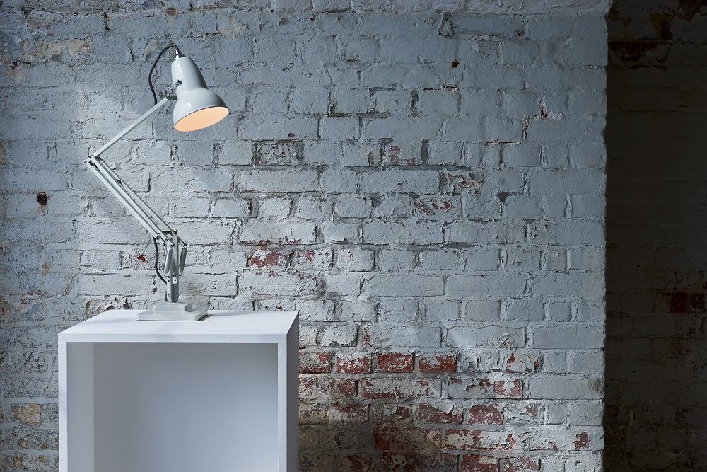 Anglepoise Original 1227 Tischleuchte in Taubengrau auf einem weißen Würfel vor einer Ziegelwand.