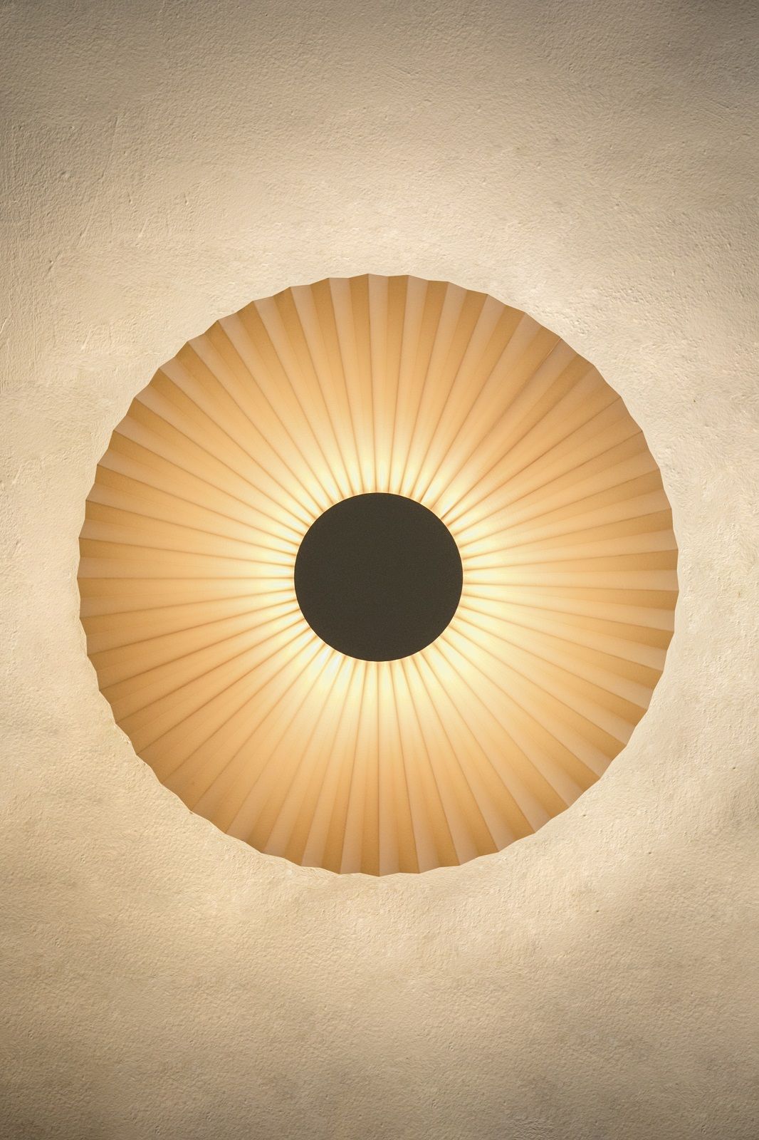 Carmen Wandleuchte von Hartô: Runde, plissierte Wandlampe mit warmem Licht und schwarzem Mittelpunkt.