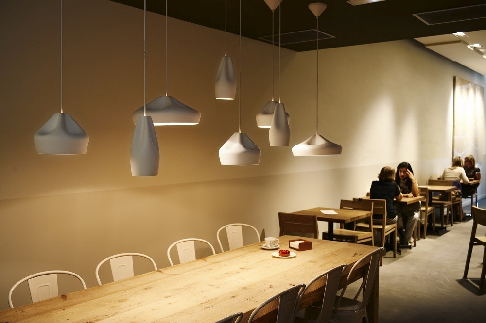 Pleat Box Pendelleuchten in Grau und Gold über einem langen Holztisch in einem Restaurant.