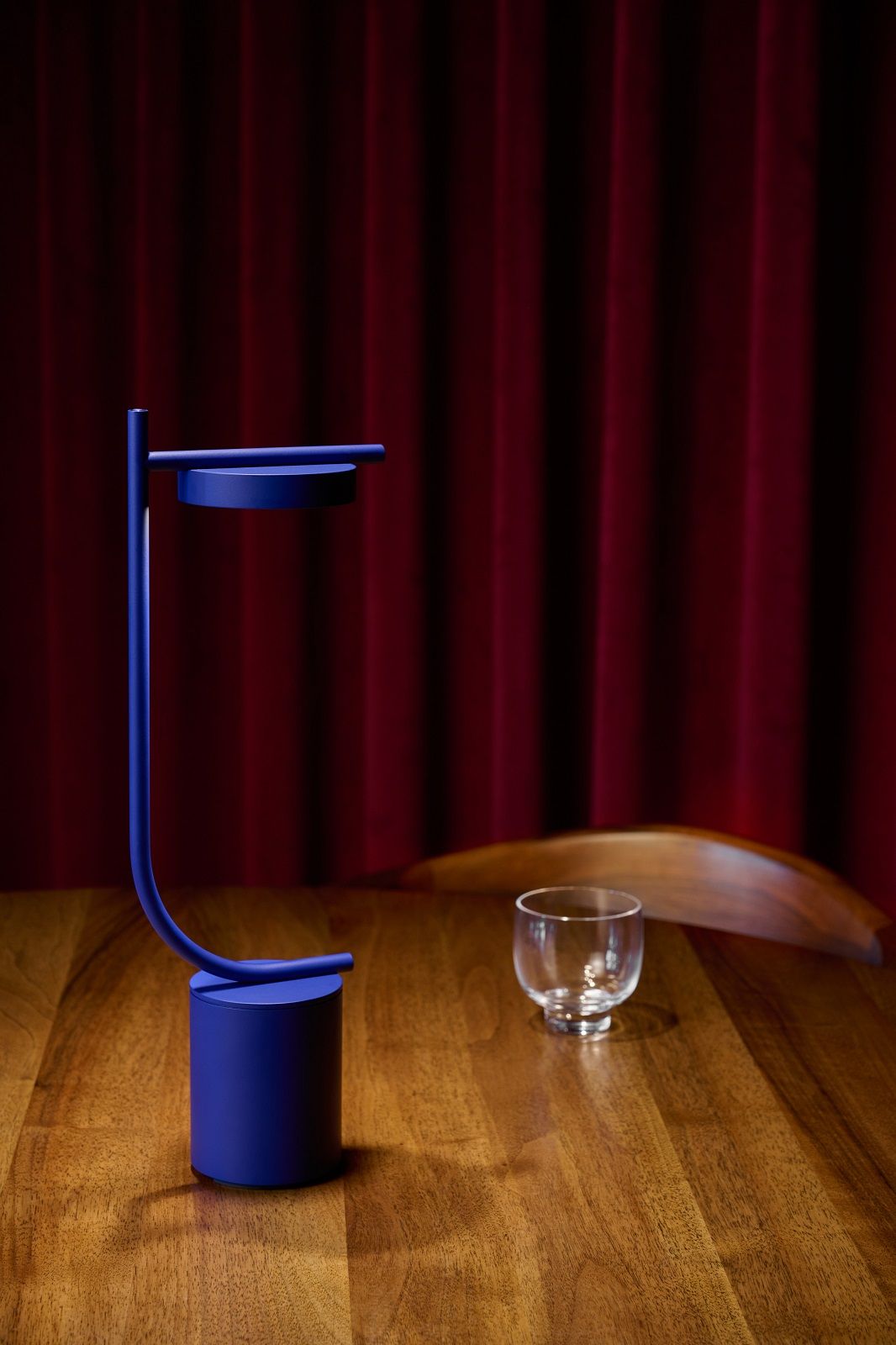Blaue Igram Tischleuchte von Grupa auf Holztisch vor rotem Vorhang. Akku-betriebene Lampe.