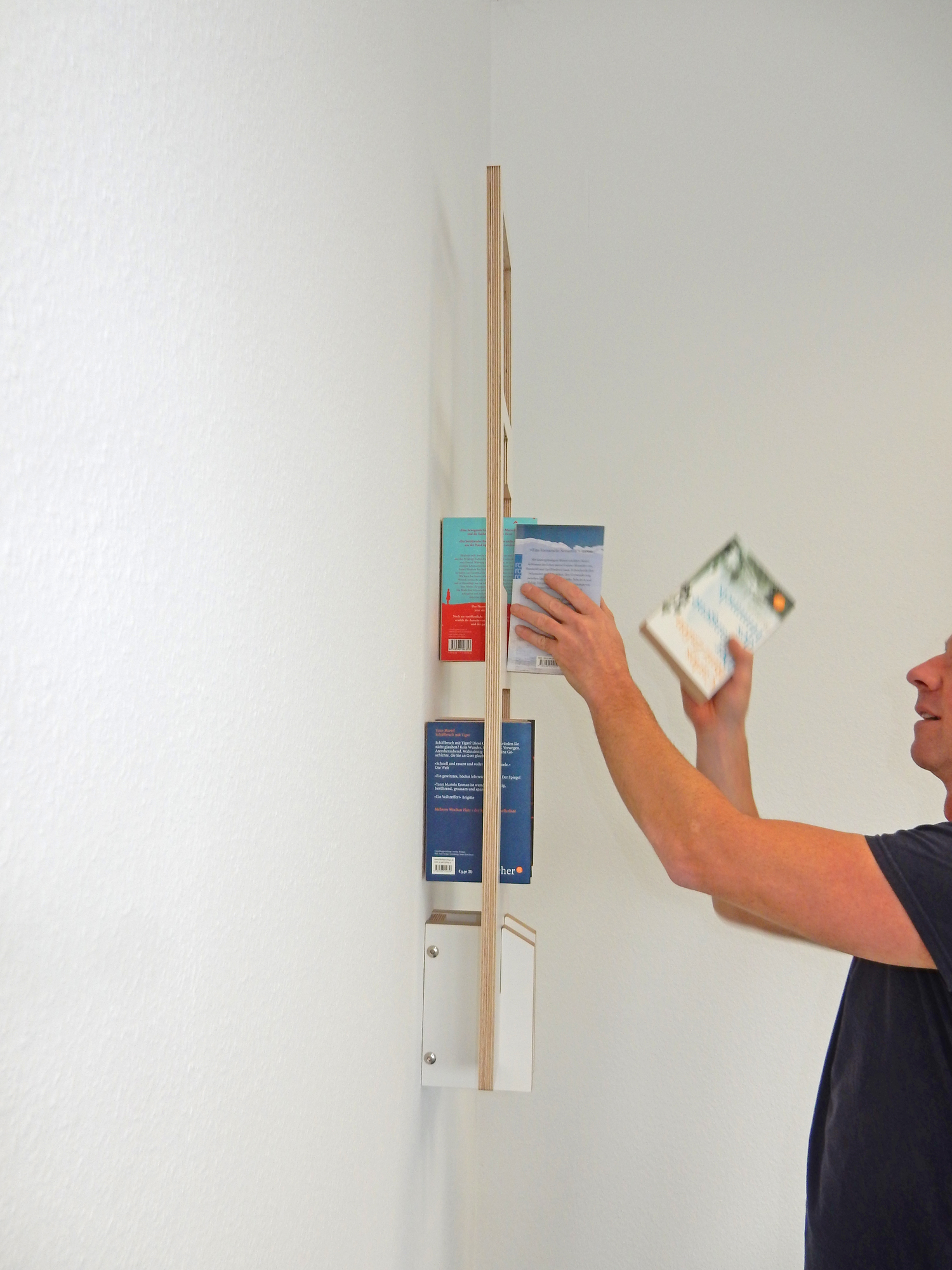 Mann bestückt ein Tojo Lesestoff Bücherregal aus Holz mit Büchern an einer weißen Wand.