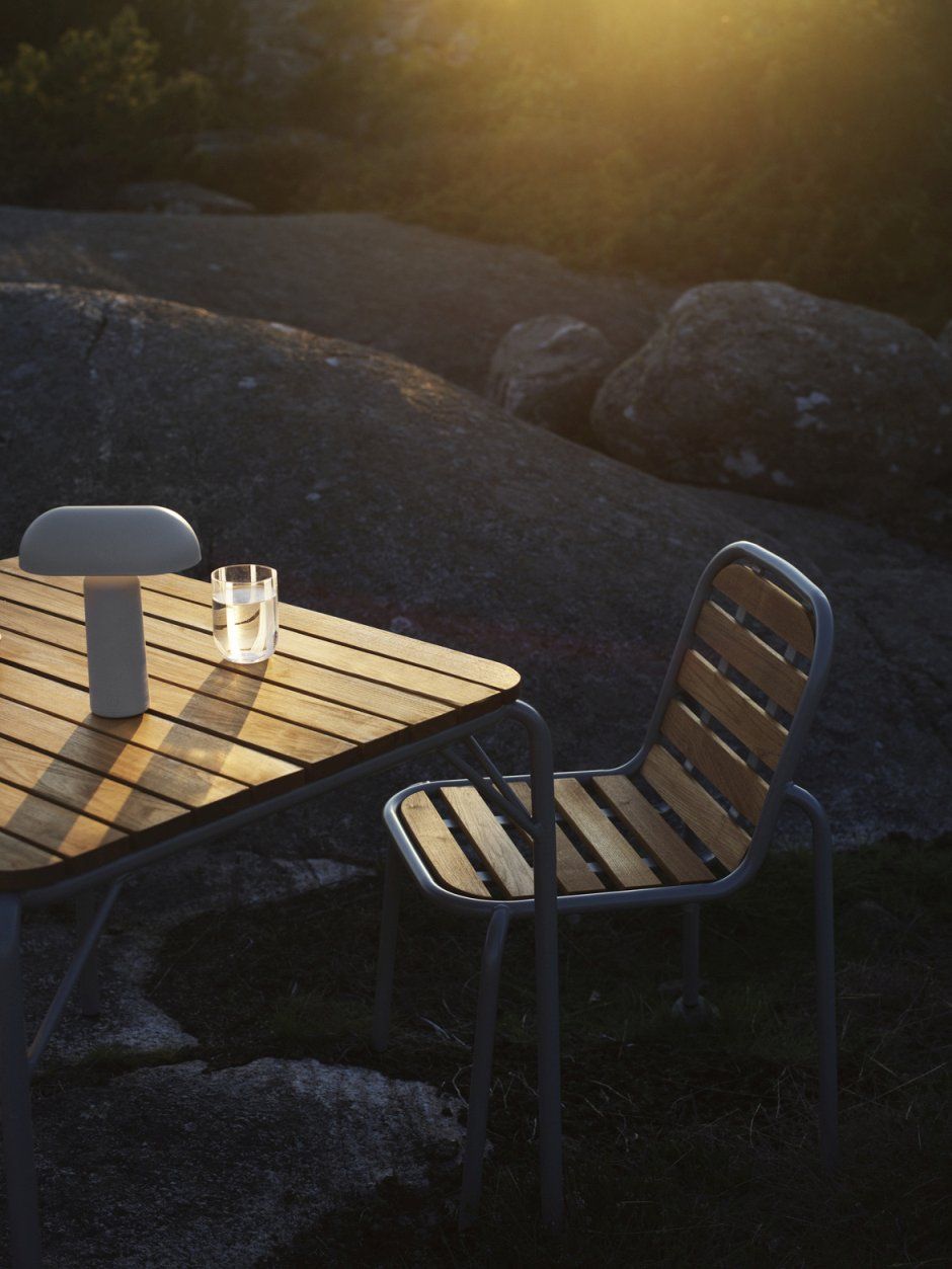 Normann Copenhagen Vig Chair Wood Stuhl Outdoor neben Tisch mit Lampe und Wasserglas im Abendlicht.