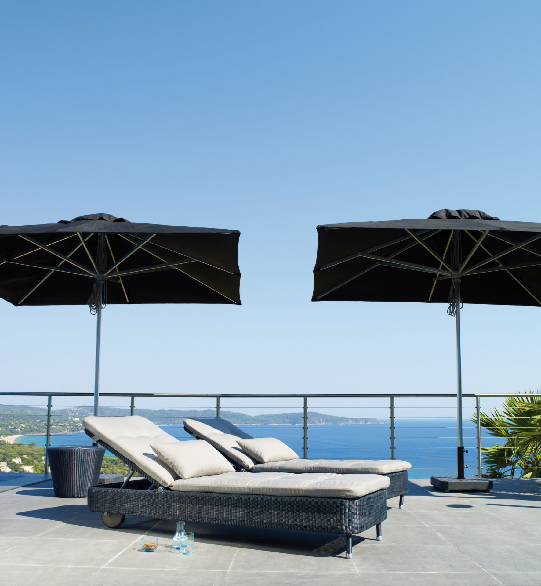 Zwei Cane-Line Sonnenliegen "Presley" in Taupe mit Kissen und Sonnenschirmen auf einer Terrasse mit Meerblick.