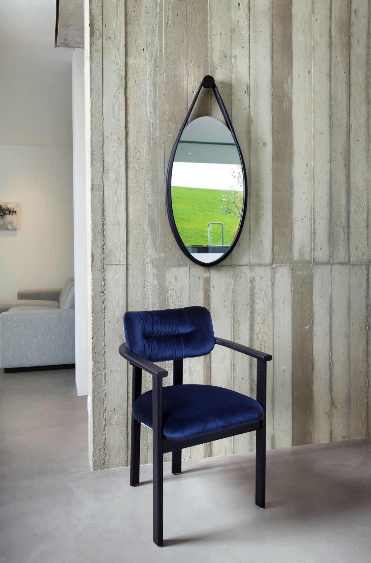 Hide Wandspiegel in Schwarz Eiche gebeizt über einem blauen Sessel vor einer Betonwand.