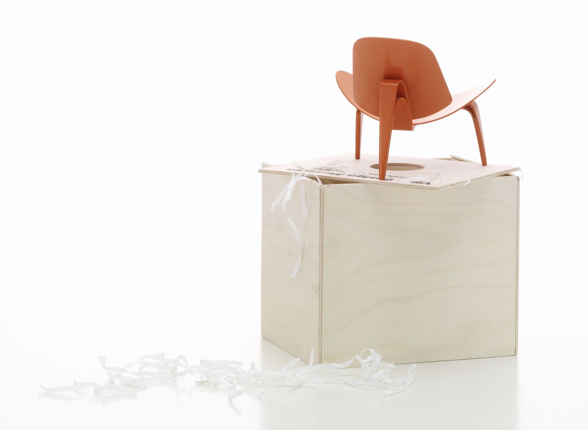 Orangefarbener 3-benet Skalstol Miniatur Stuhl von Vitra auf einer Holzkiste mit Papierstreifen.