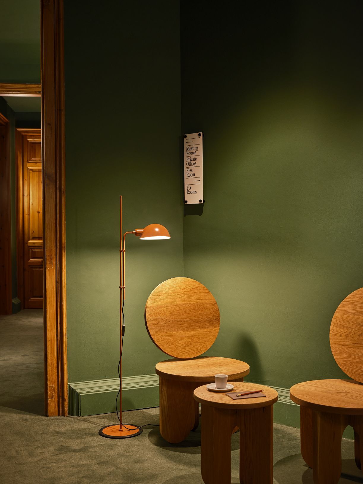 Funiculí Stehleuchte in moosgrau von Marset neben Holztischen und grüner Wand.