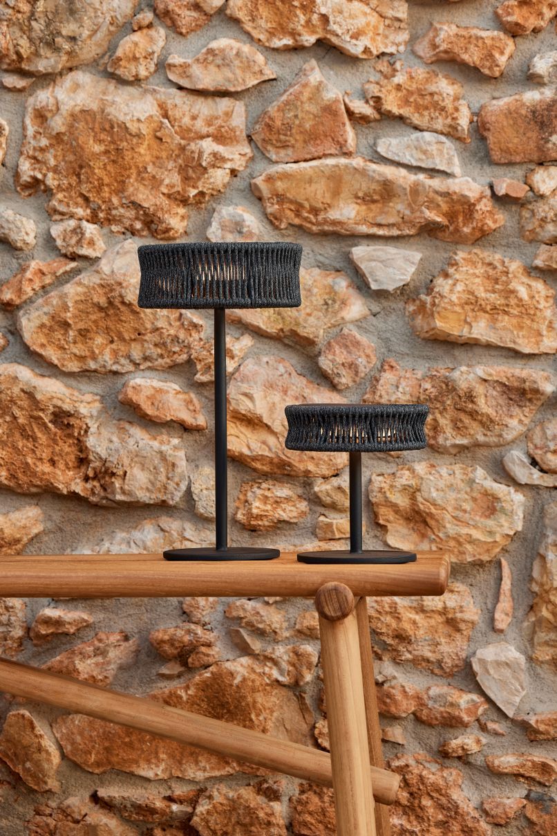 Zwei Cane-Line Illusion Outdoor Tischleuchten in Taupe auf einem Holztisch vor Steinwand.