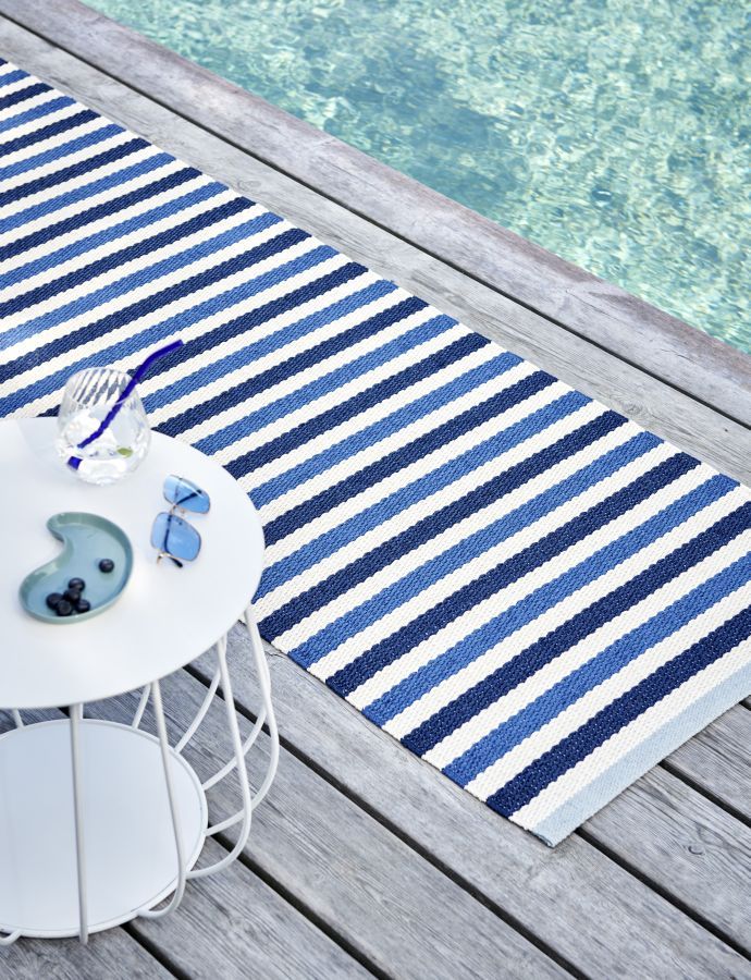 Pappelina Teo Teppich Denim gestreift auf Holzdeck neben Pool, mit Tisch und Accessoires.