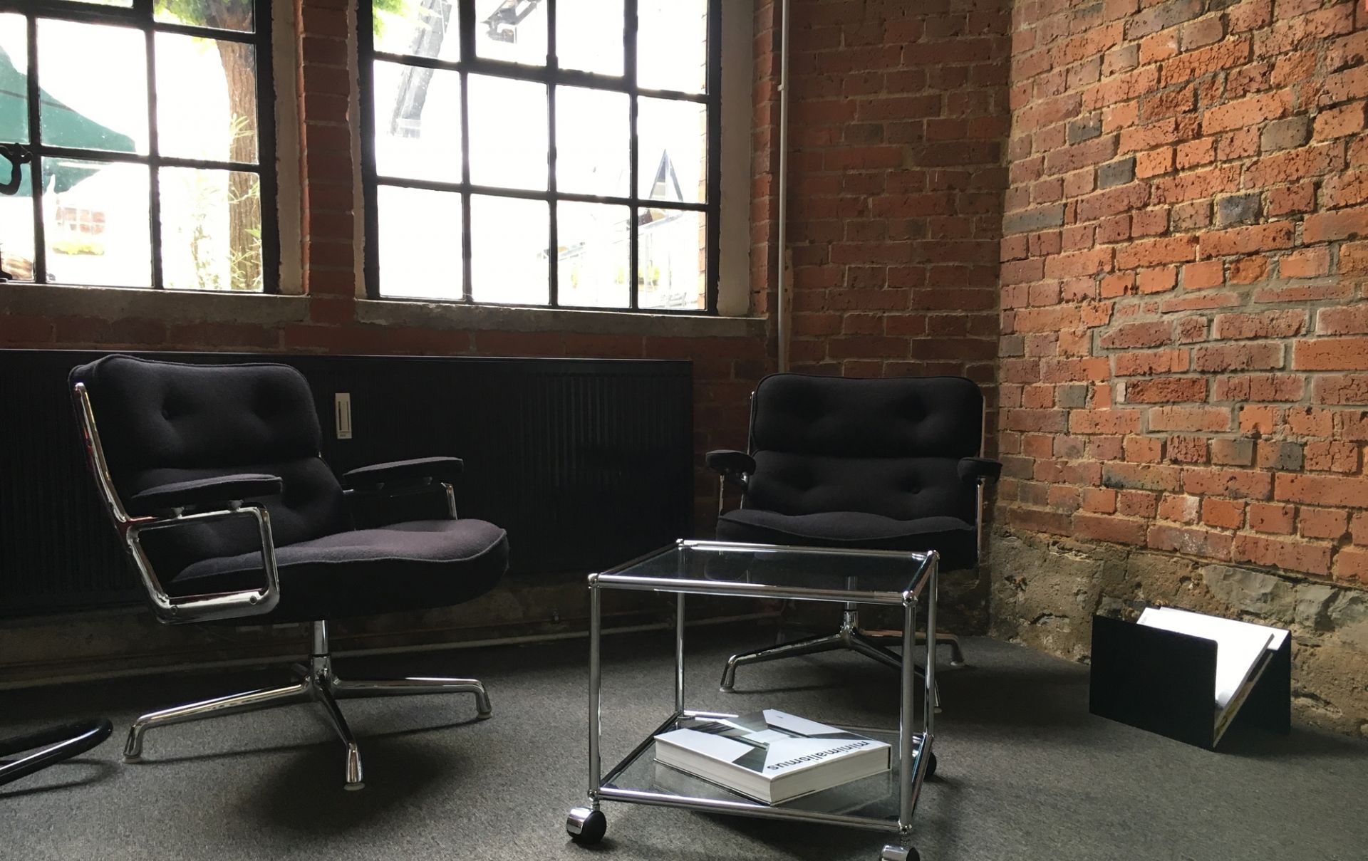 ZS01 M Stadie Zeitungsständer aus Metall neben schwarzen Sesseln und Glastisch in Loft-Atmosphäre.