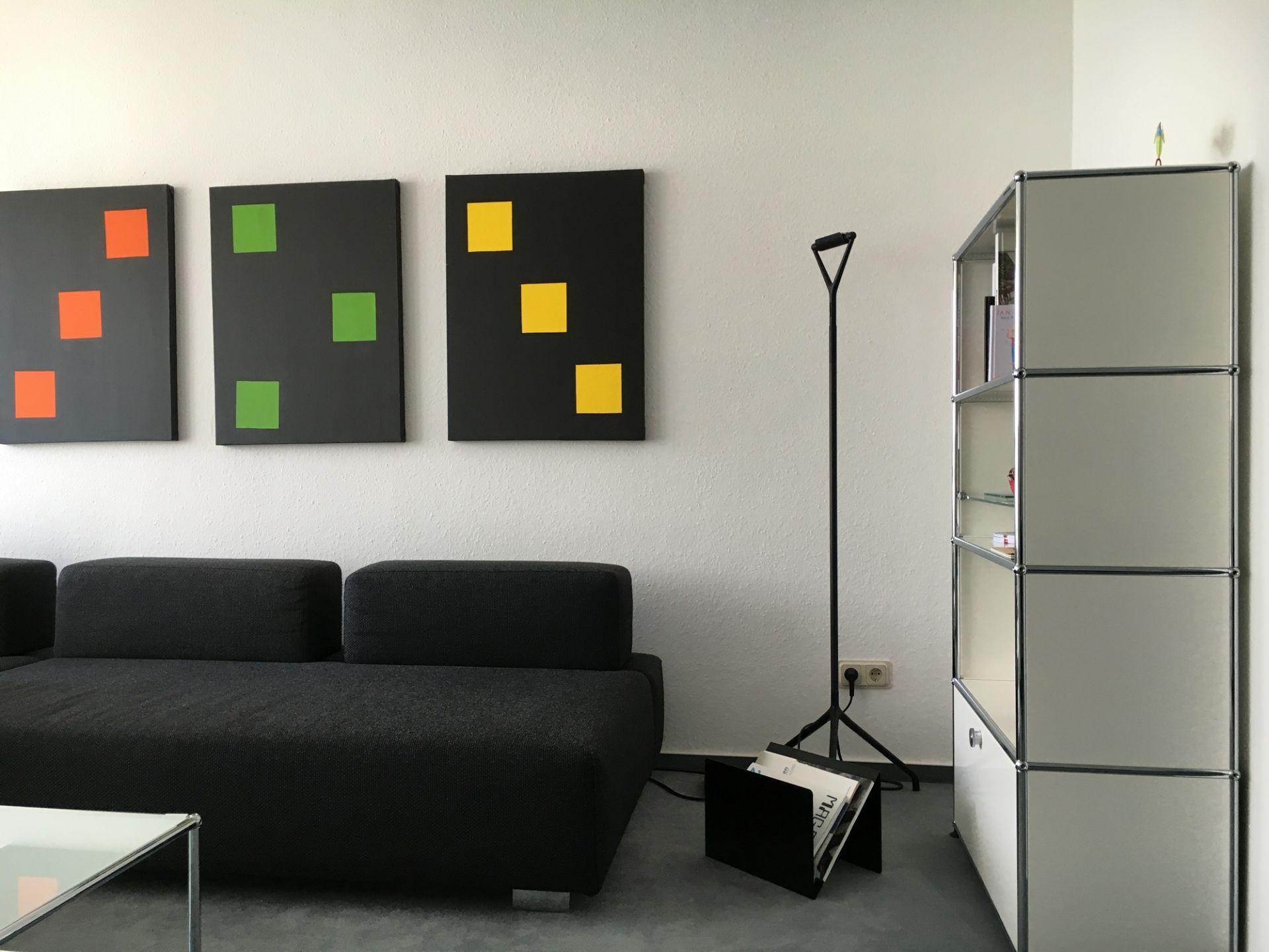 Schwarzer ZS01 M Zeitungsständer Stadie aus Metall neben einer Stehlampe und einem Sofa im Wohnzimmer.