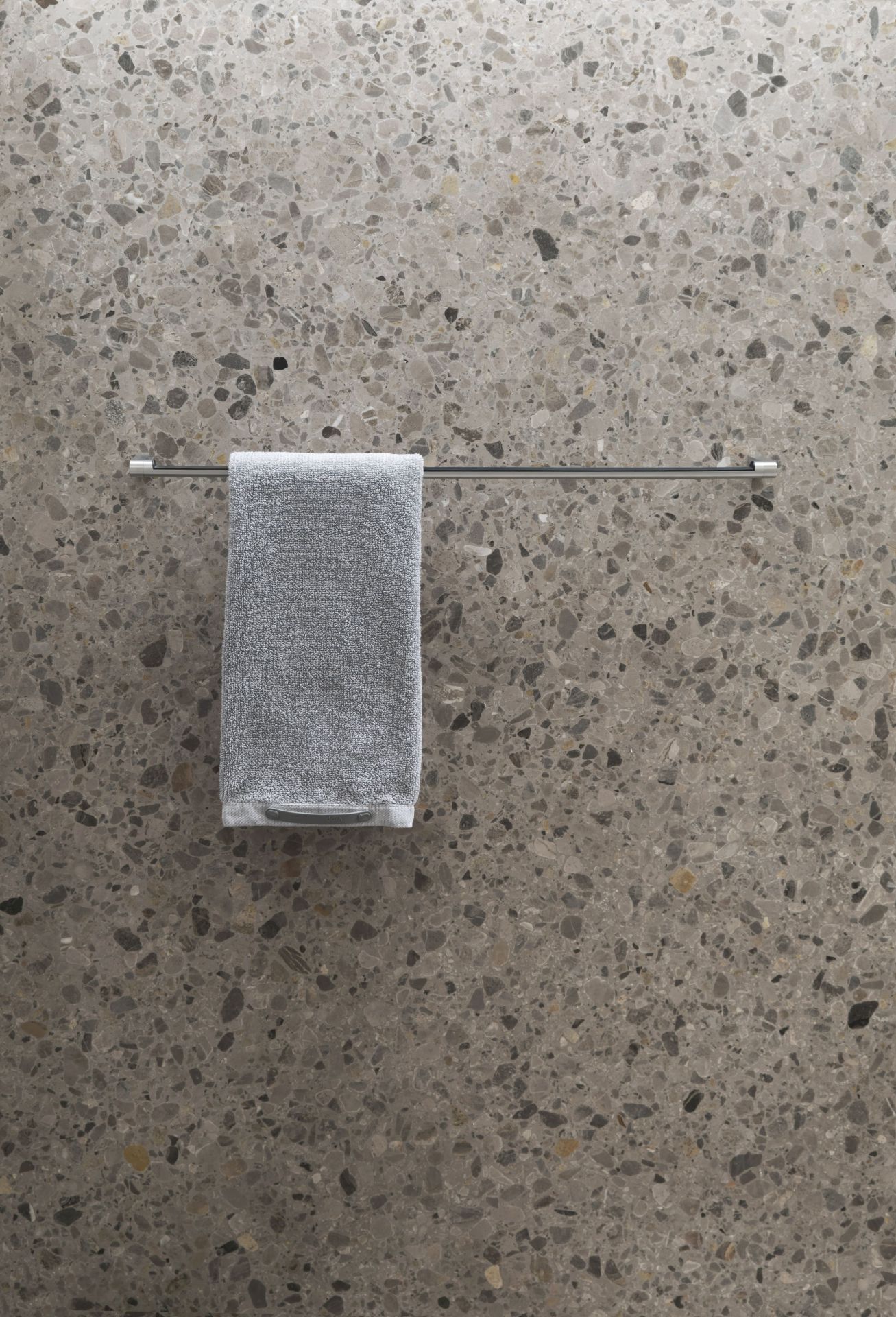 Silberne Vipp Handtuchstange mit grauem Handtuch vor einer grauen Wand mit Steinmuster.