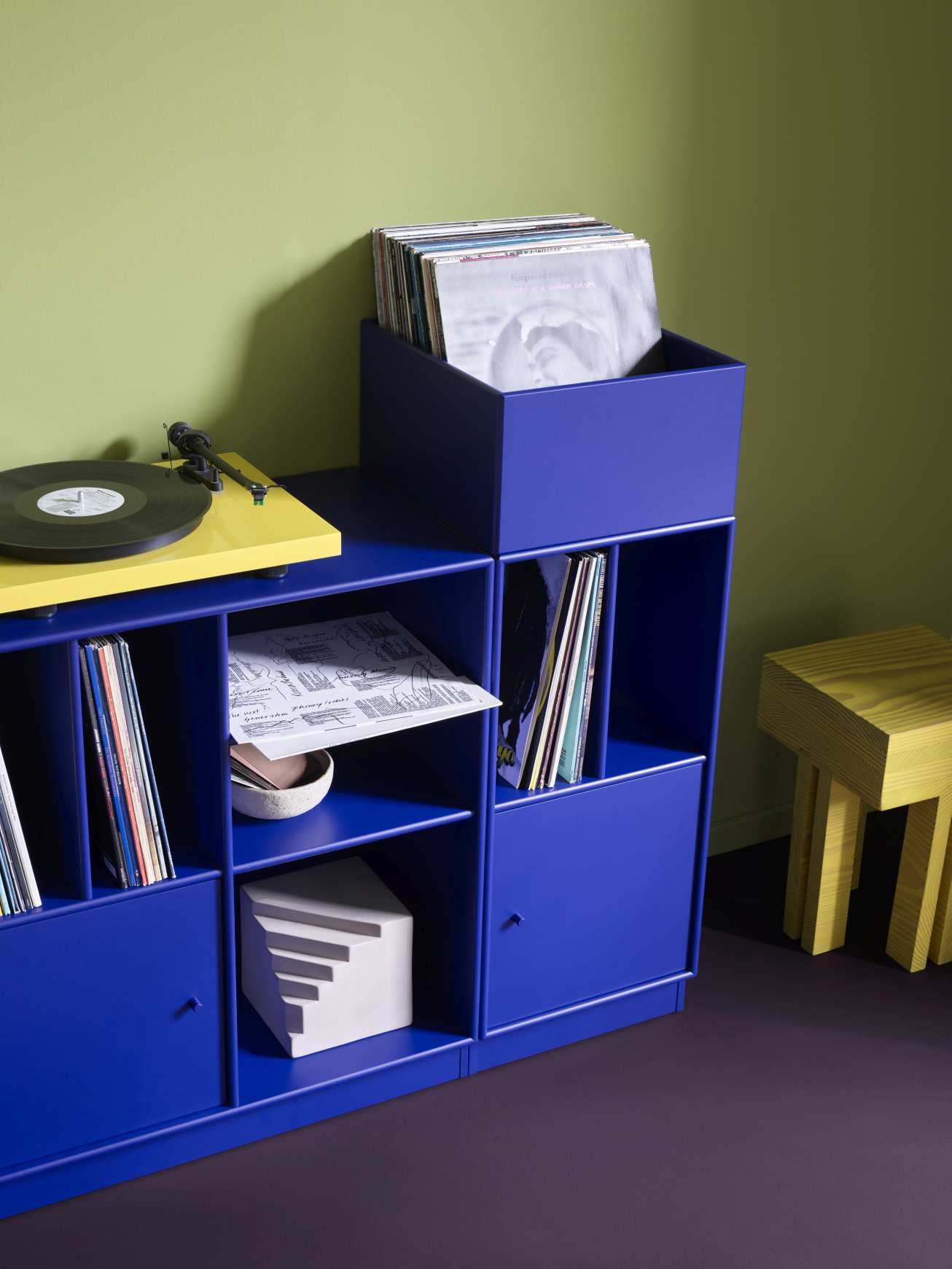 Blaues Spin III Schallplattenregal mit Schallplatten, Plattenspieler und gelbem Hocker für moderne Vinyl-Aufbewahrung.