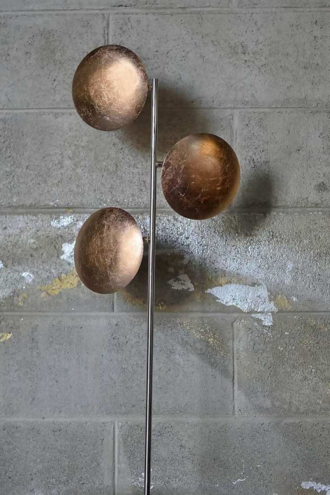 Lederam Stehleuchte mit drei goldenen Kugeln vor einer grauen Ziegelwand.
