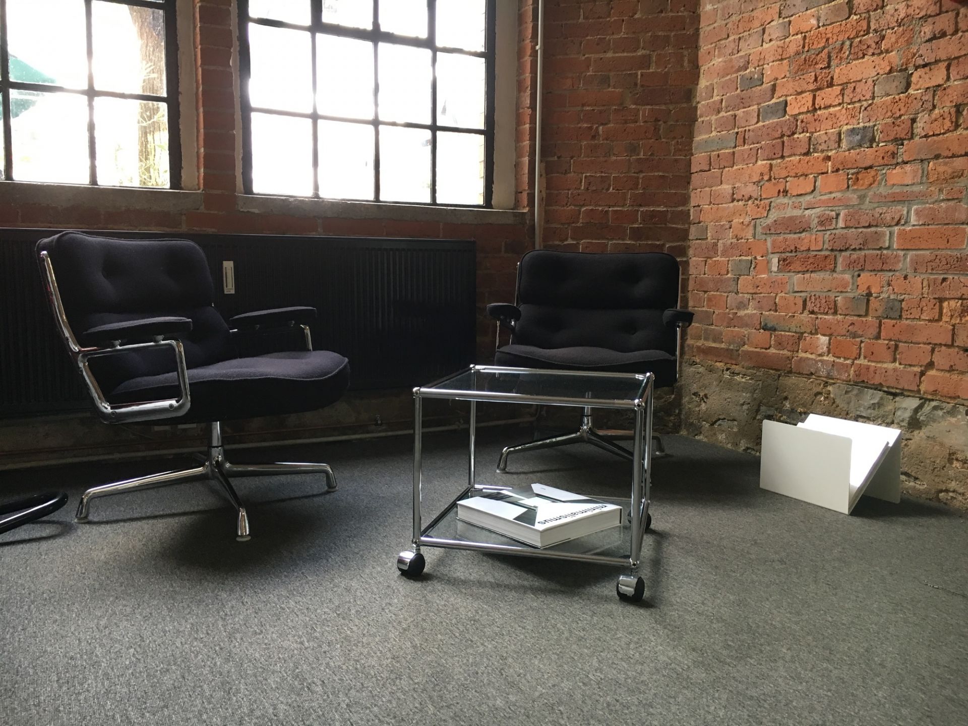 ZS01 M Zeitungsständer Stadie aus Glas und Chrom mit Rollen, neben zwei Bürostühlen.