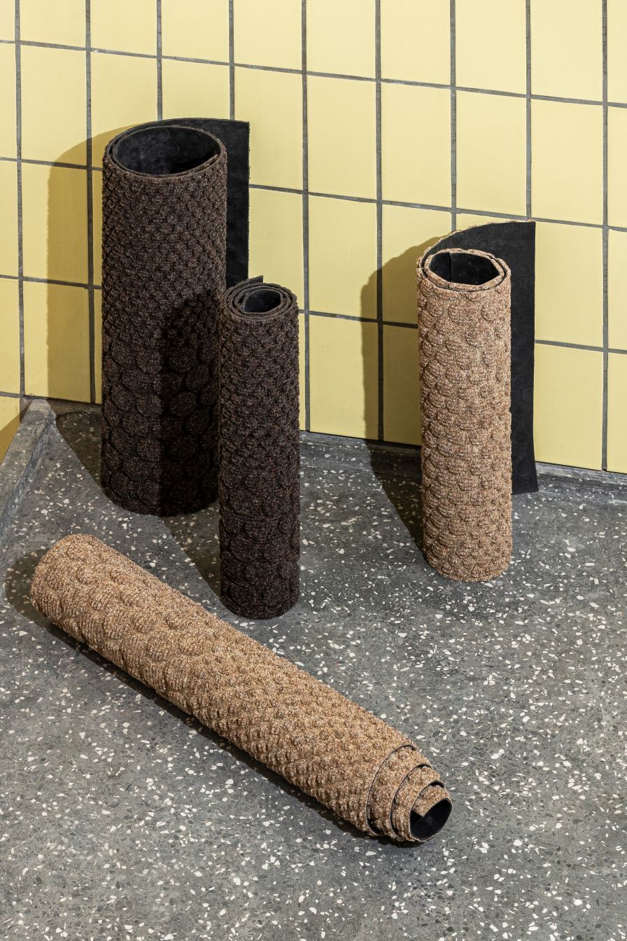 Hex Fußmatte Heymat: Gerollte Fußmatten in Braun- und Beigetönen auf grauem Untergrund, moderne Eingangsmatte.