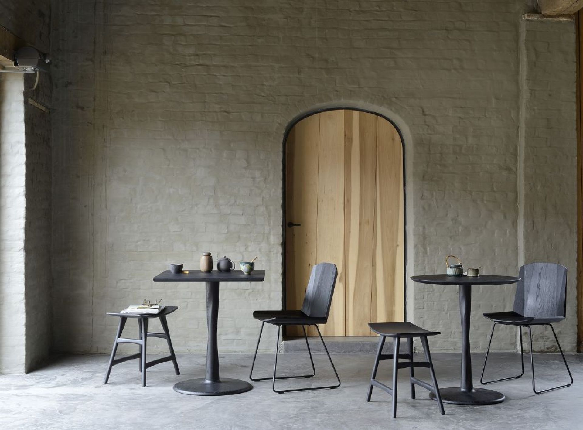 Ethnicraft Torsion Eiche Esstisch B 70 schwarz mit passenden Stühlen und Hockern vor Backsteinwand.