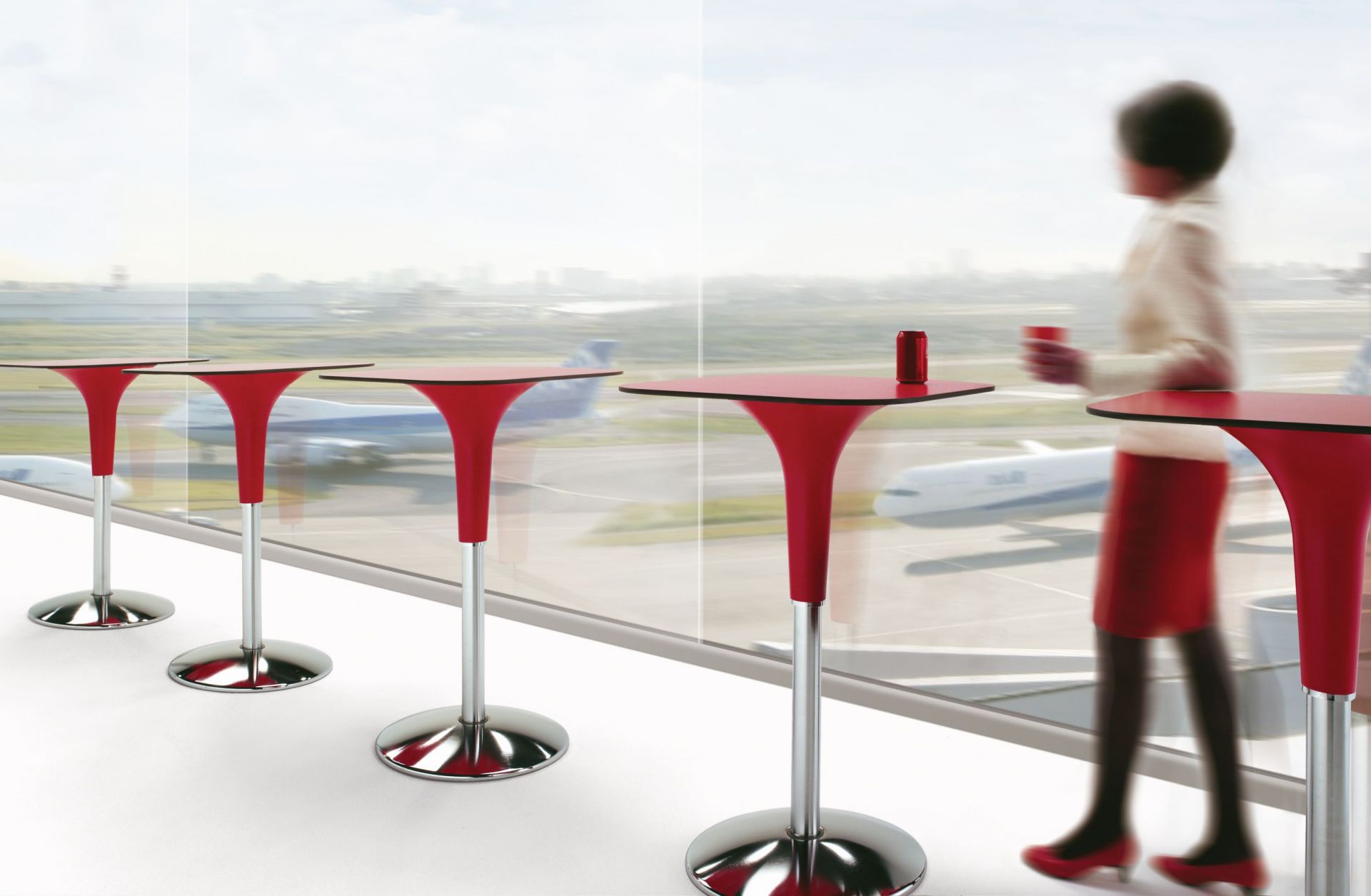 Zanziplano Tisch von Rexite, roter Stehtisch mit Frau und Getränk vor Fenster mit Flugzeugen.