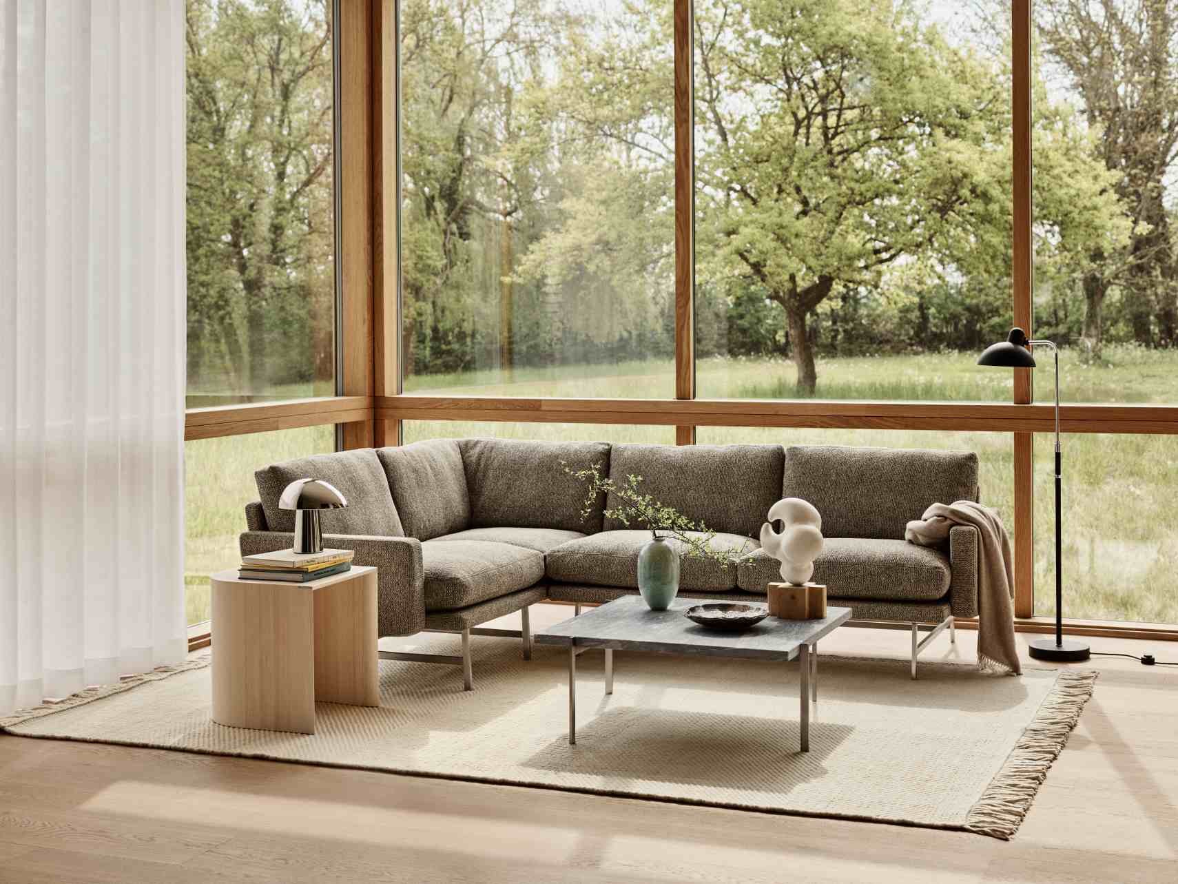 Wohnzimmer mit Fritz Hansen Hocker/Beistelltisch aus Holz und grauem Ecksofa vor großem Fenster.