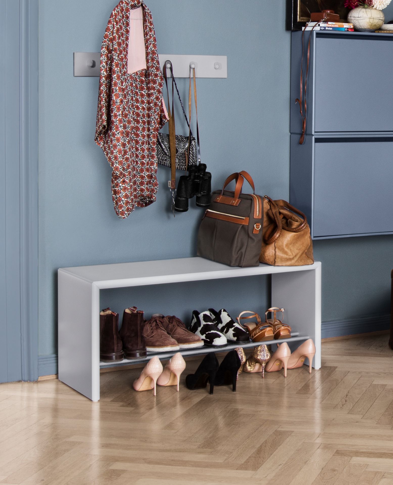 Helle Schuhbank mit zwei Ablagen, gefüllt mit Schuhen. Darüber Taschen und Jacke an der Wand.