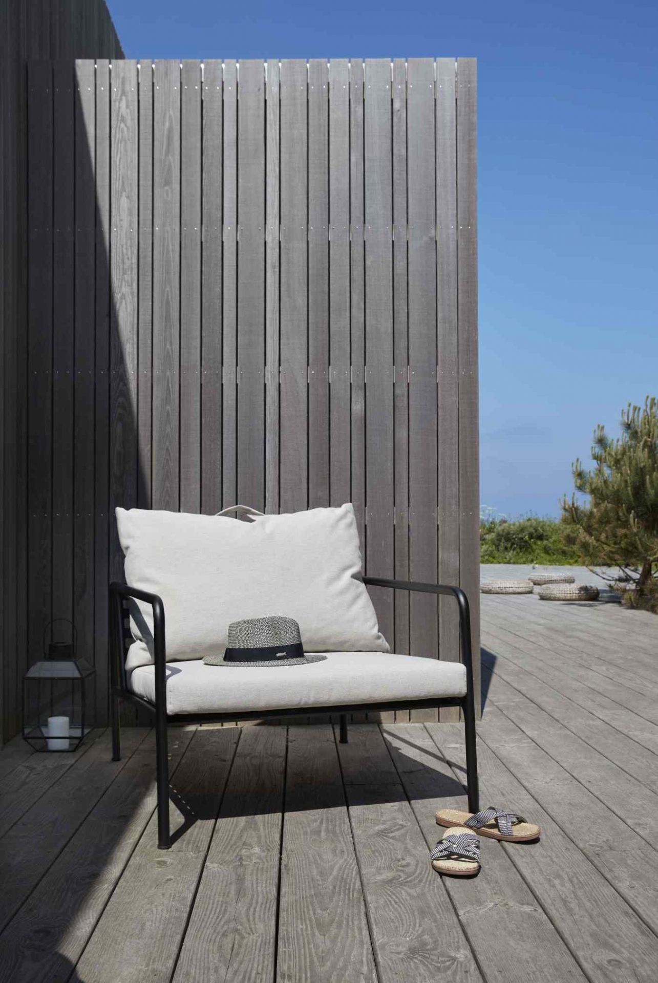 Avon Lounge Chair von Houe: Bequemer Outdoor Sessel mit Hut und Sandalen auf Holzterrasse.
