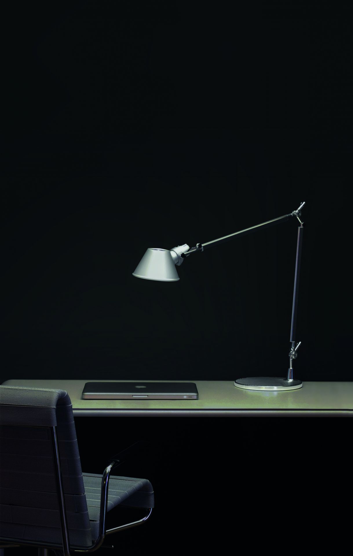 Silberne Tolomeo LED Tischleuchte von Artemide auf Schreibtisch mit Laptop und Bürostuhl vor schwarzem Hintergrund.