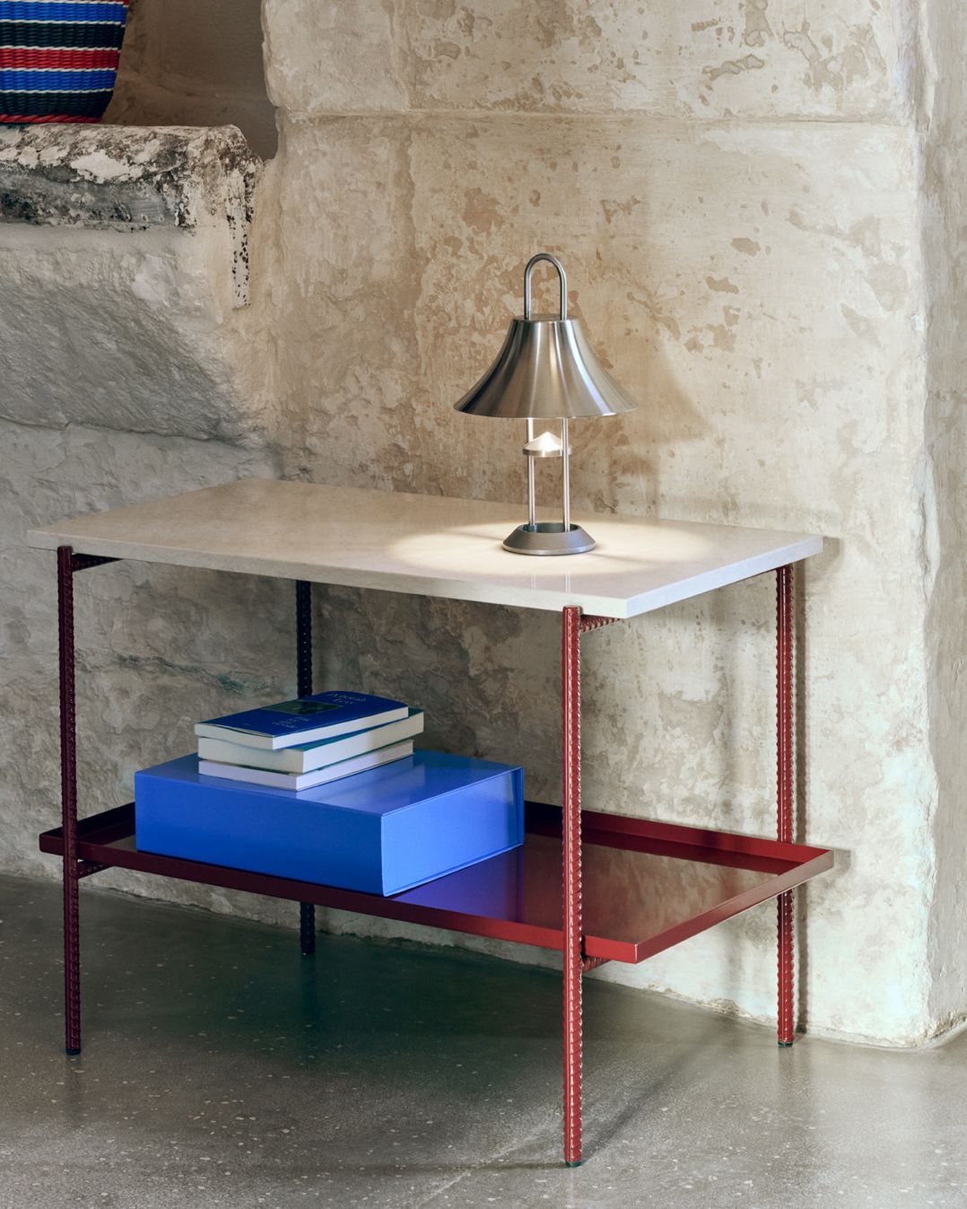 Roter Hay Rebar Beistelltisch mit Ablage, Bücher und Lampe vor einer Steinwand.