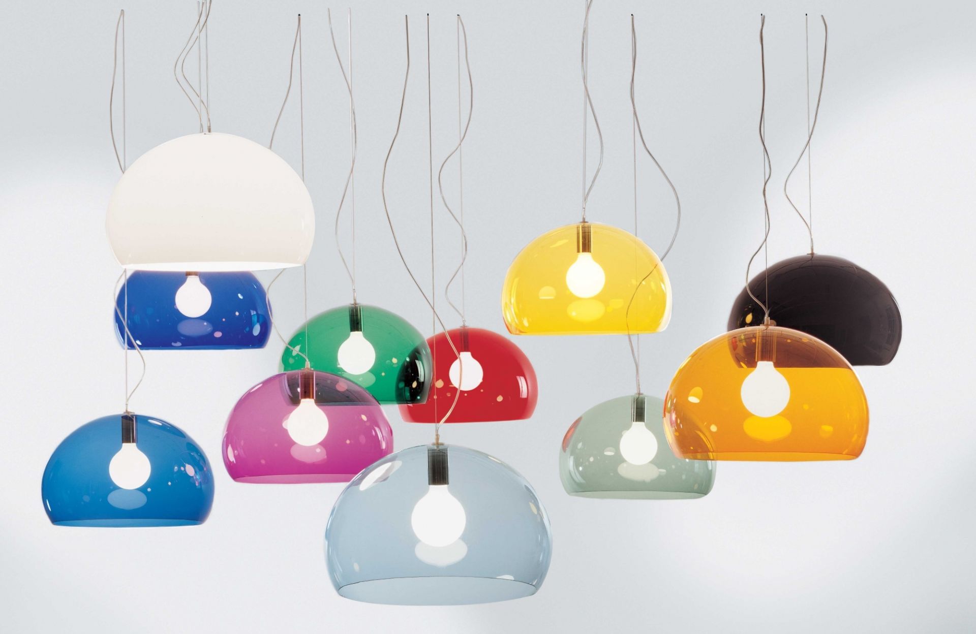 Mehrere farbige Fly Pendelleuchten von Kartell, darunter wolkenblau transparent, hängen vor einem weißen Hintergrund.