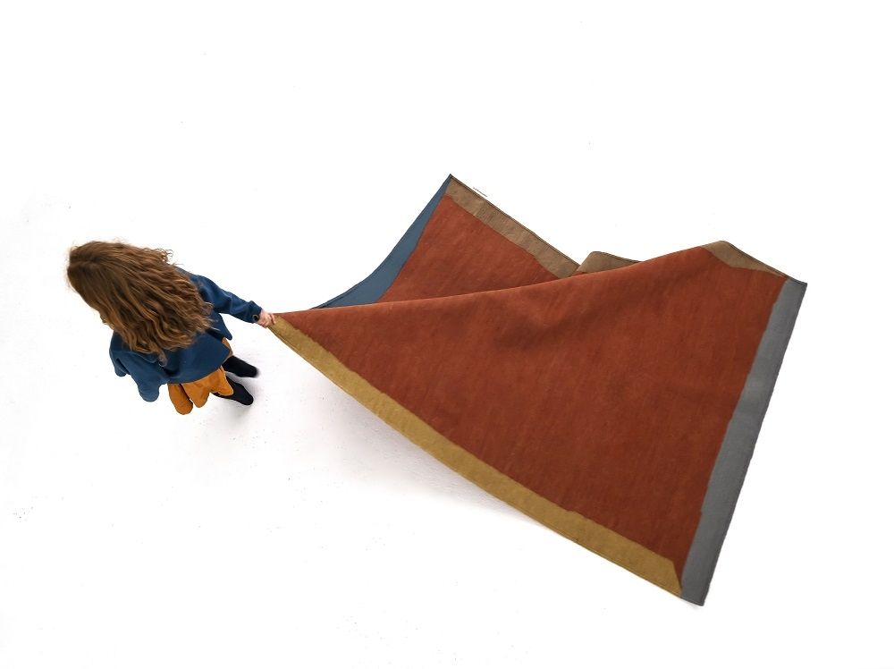 Mädchen hält Nanimarquina Tones Kilim Teppich in Rotbraun, Blau und Grau auf weißem Hintergrund.