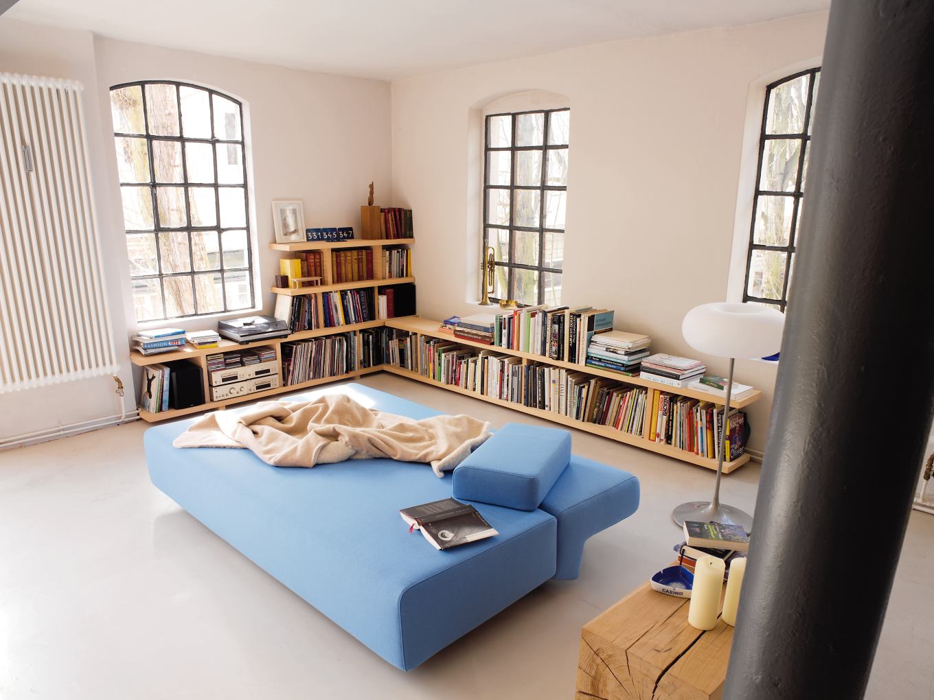 Helles Wohnzimmer mit blauem Cosma Schlafsofa, Bücherregal und großen Fenstern. Gemütliches Schlafsofa für modernes Wohnen.