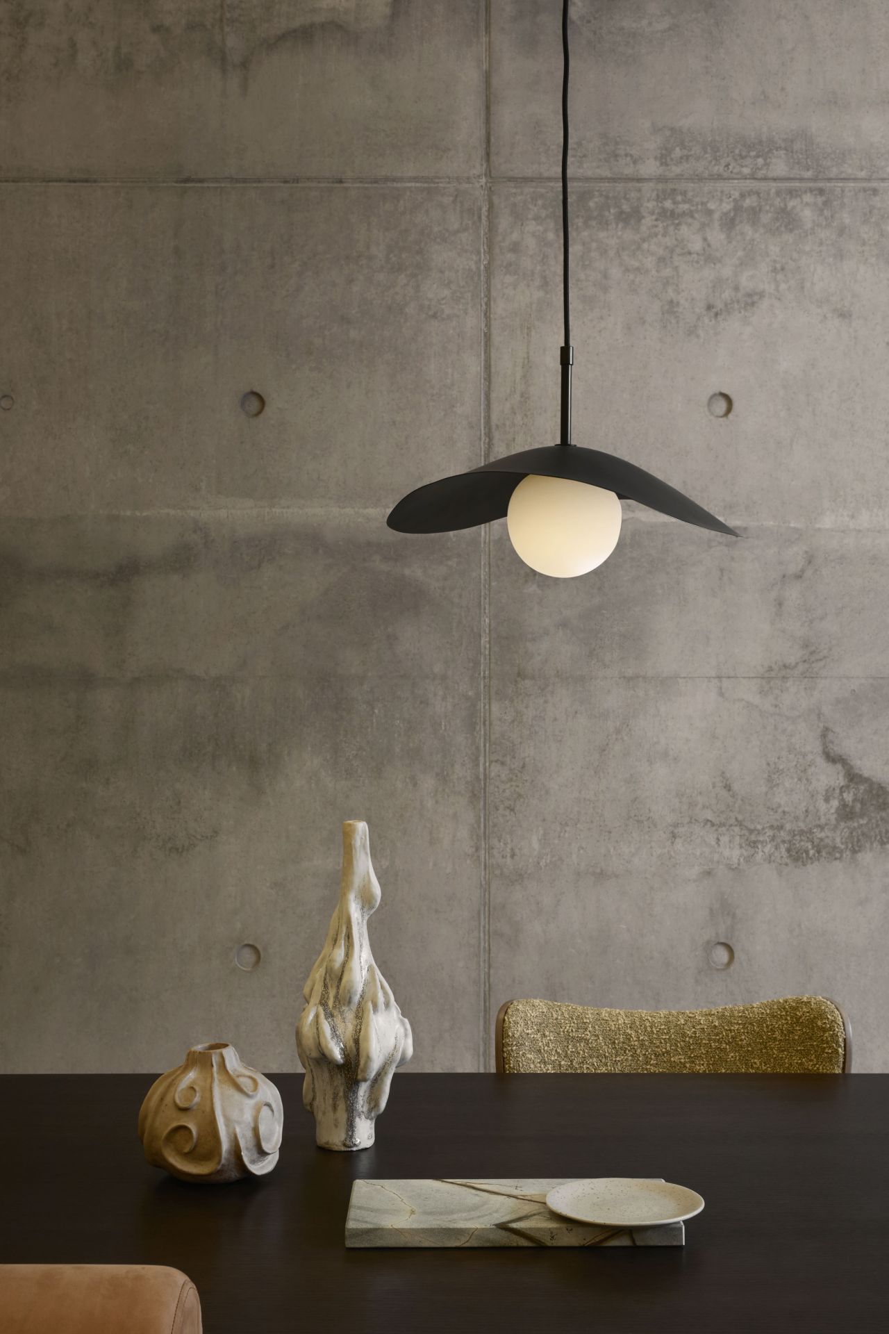 Fuji Pendelleuchte von Norr11 über Esstisch mit Deko-Objekten vor Betonwand. Design Lampe.