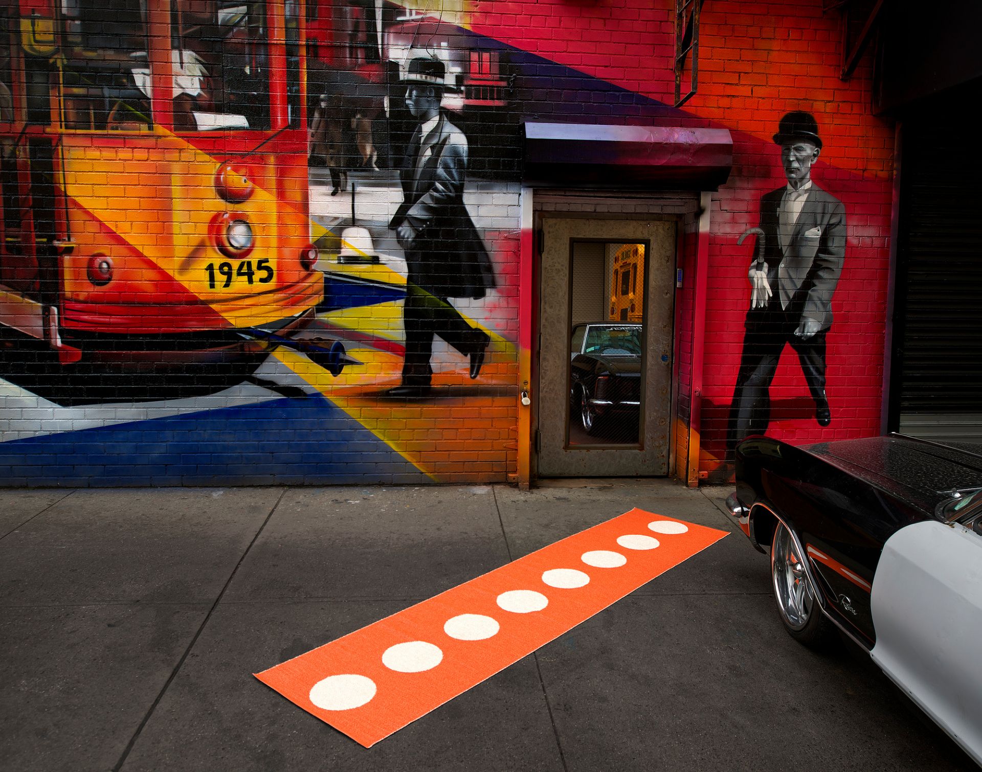 Vera Kunststoffteppich von Pappelina, orangefarbener Läufer mit weißen Punkten vor einer bemalten Hauswand in einer Großstadt.