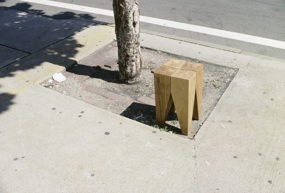 E15 Backenzahn Hocker aus Holz neben einem Baum auf einem Bürgersteig.
