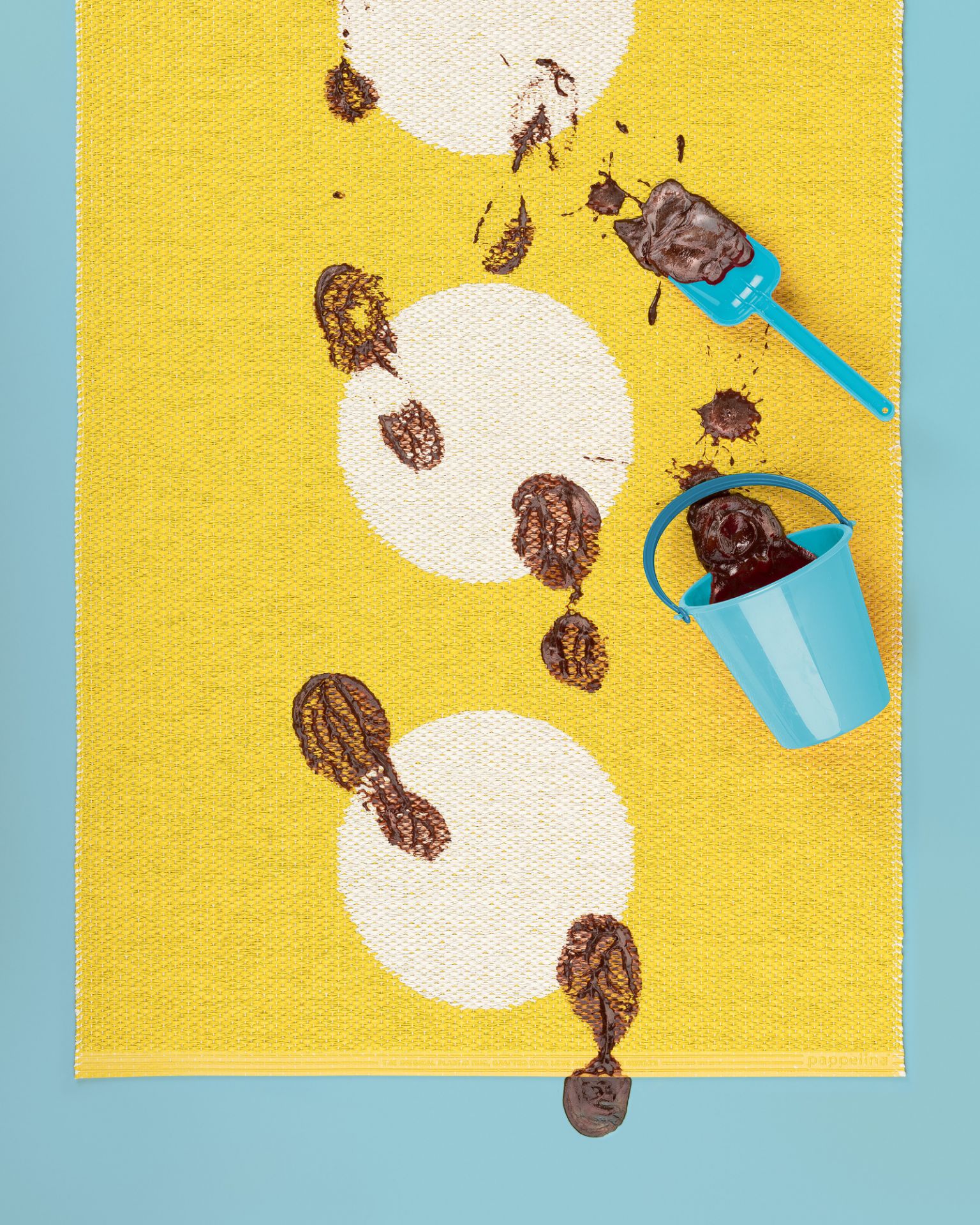 Vera Kunststoffteppich Pappelina in Gelb mit weißen Kreisen, dekoriert mit Schokoladenfußabdrücken, Eimer und Schaufel.