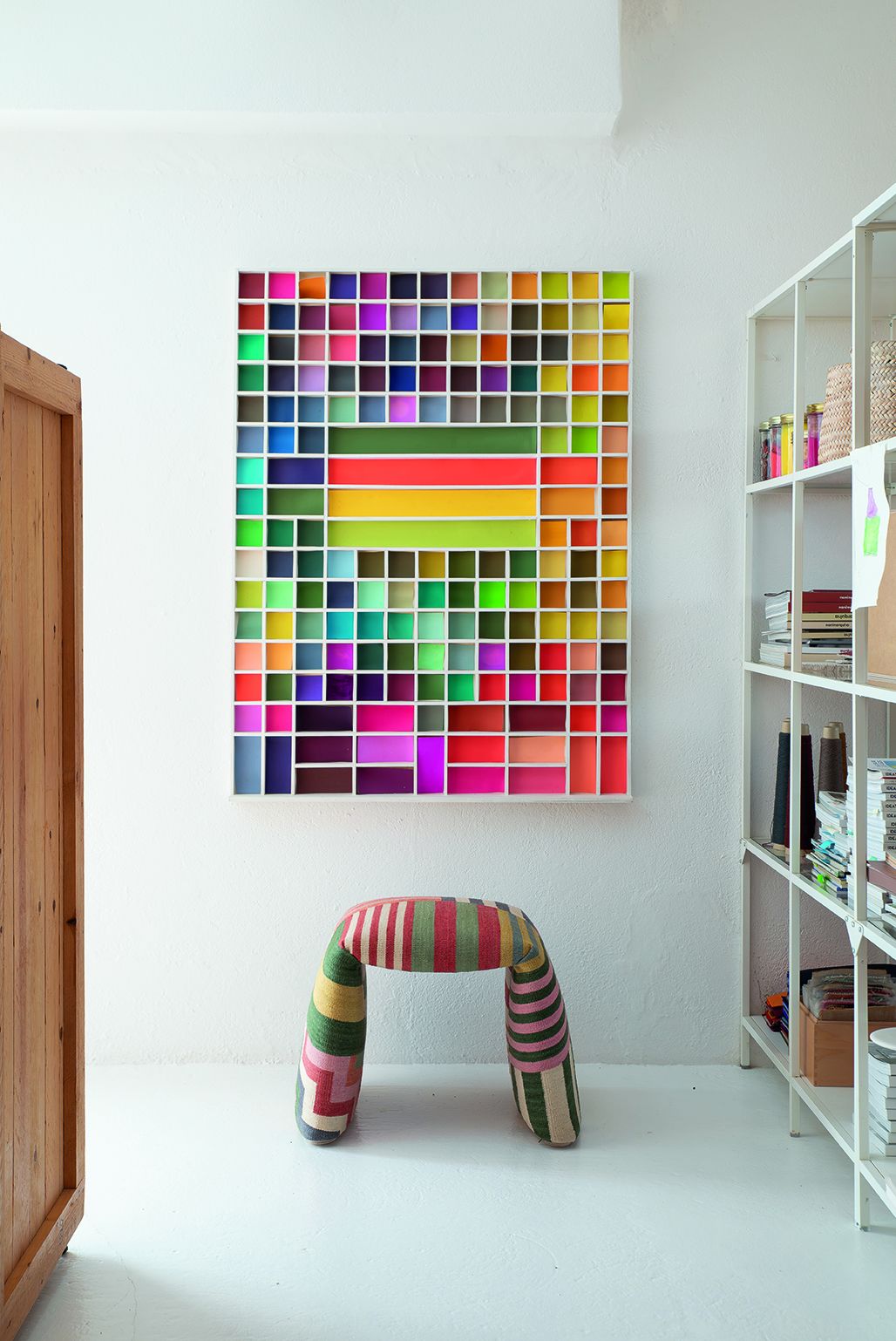 Nanimarquina Folded Pattern Bank: Gepolsterte Sitzbank mit bunten Streifen vor farbenfrohem Wandbild.