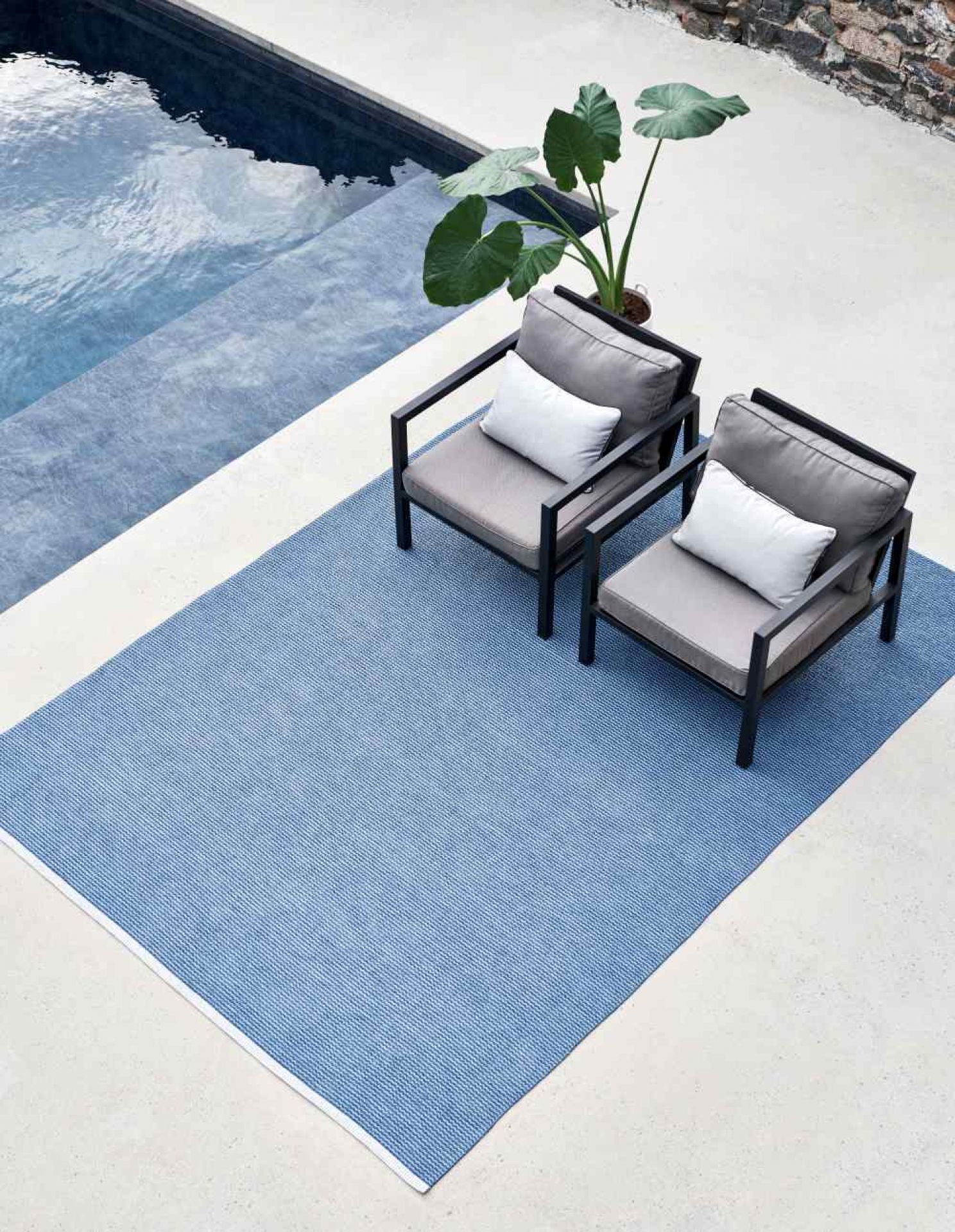 Blauer Kunststoffteppich von Pappelina (ca. 230x320 cm) im Außenbereich mit zwei Stühlen und Pflanze.