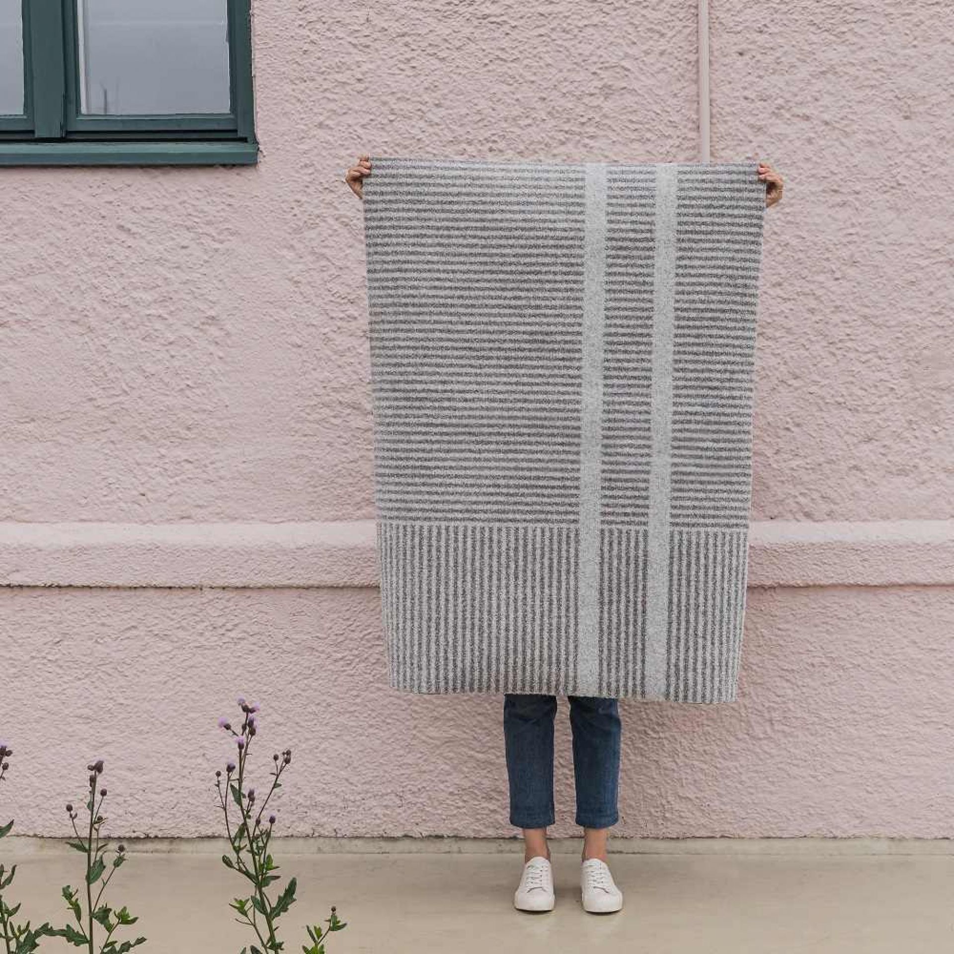 Person hält graue Løype Fußmatte von Heymat mit Streifenmuster vor einem rosa Haus.