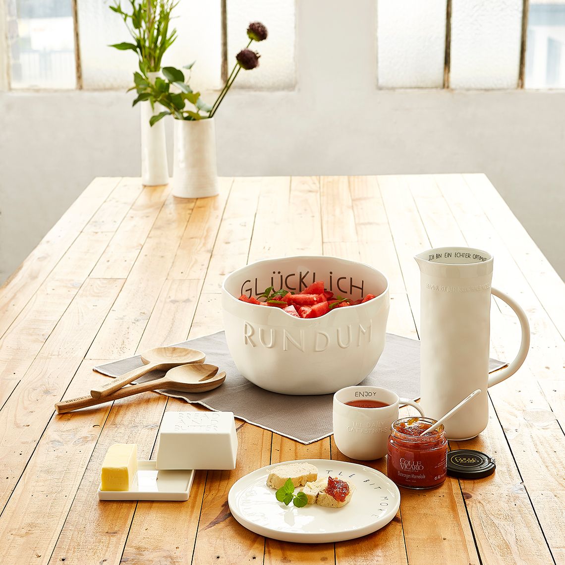 Tischgedeck mit "Ich bin ein echter Optimist" Krug, Schüssel, Marmelade und Frühstücksgeschirr für einen guten Morgen.