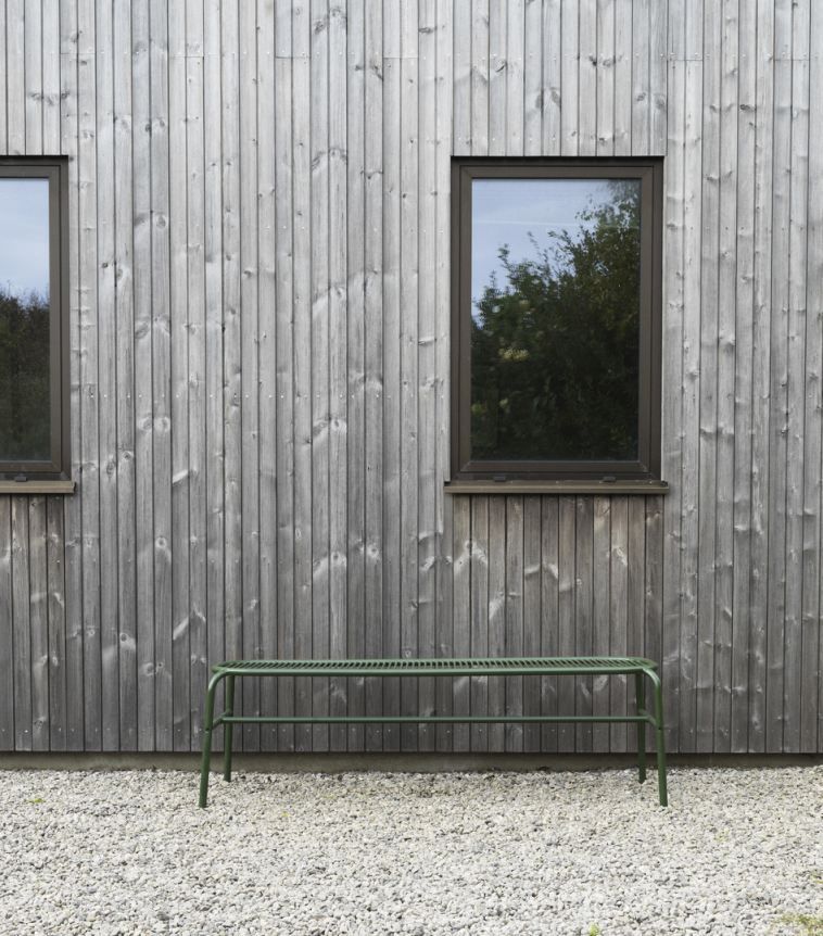 Grüne Vig Bench Bank von Normann Copenhagen für den Außenbereich vor grauer Holzwand.