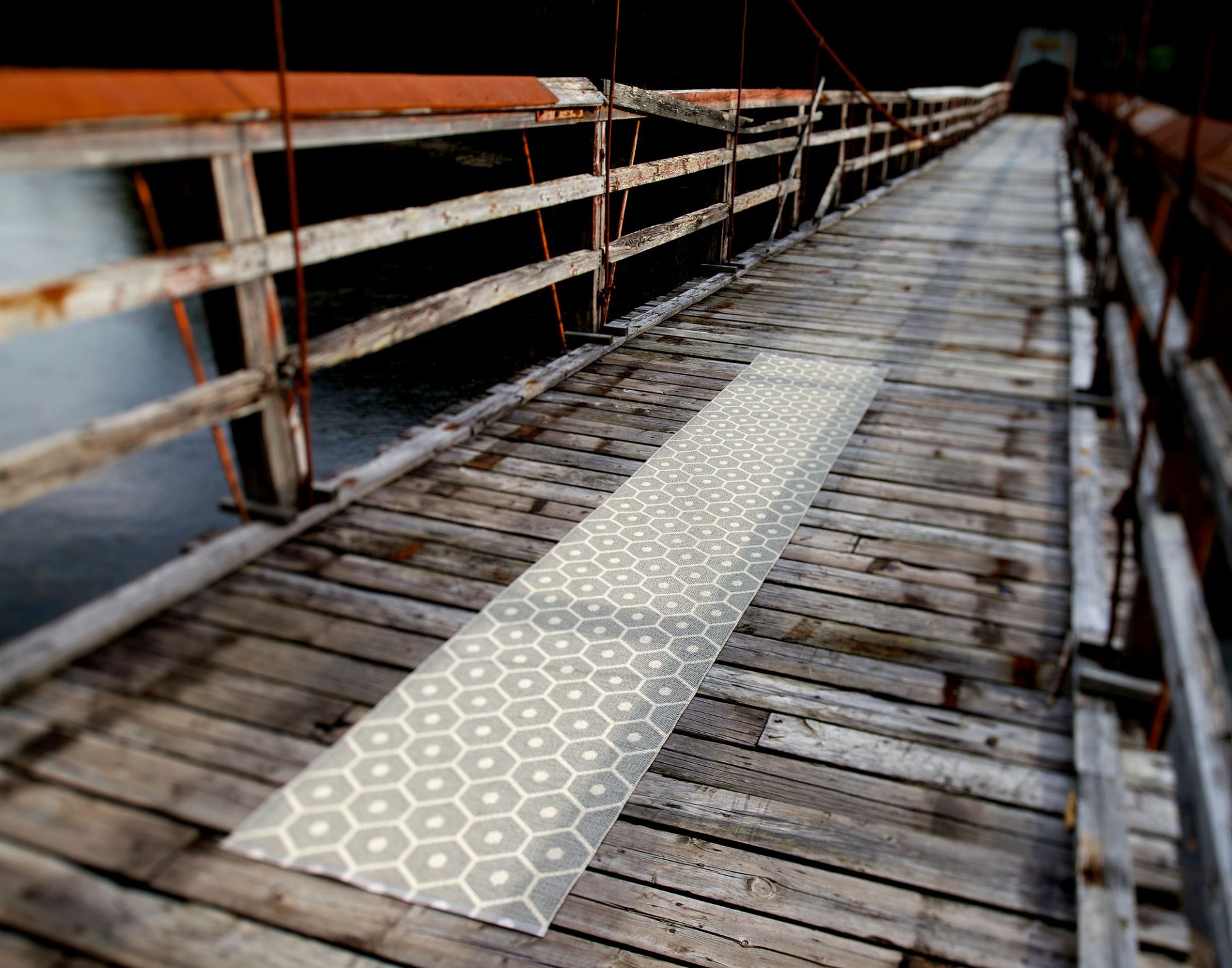 Honey Kunststoffteppich von Pappelina, 70x160 cm, mit geometrischem Muster auf einer Holzbrücke.