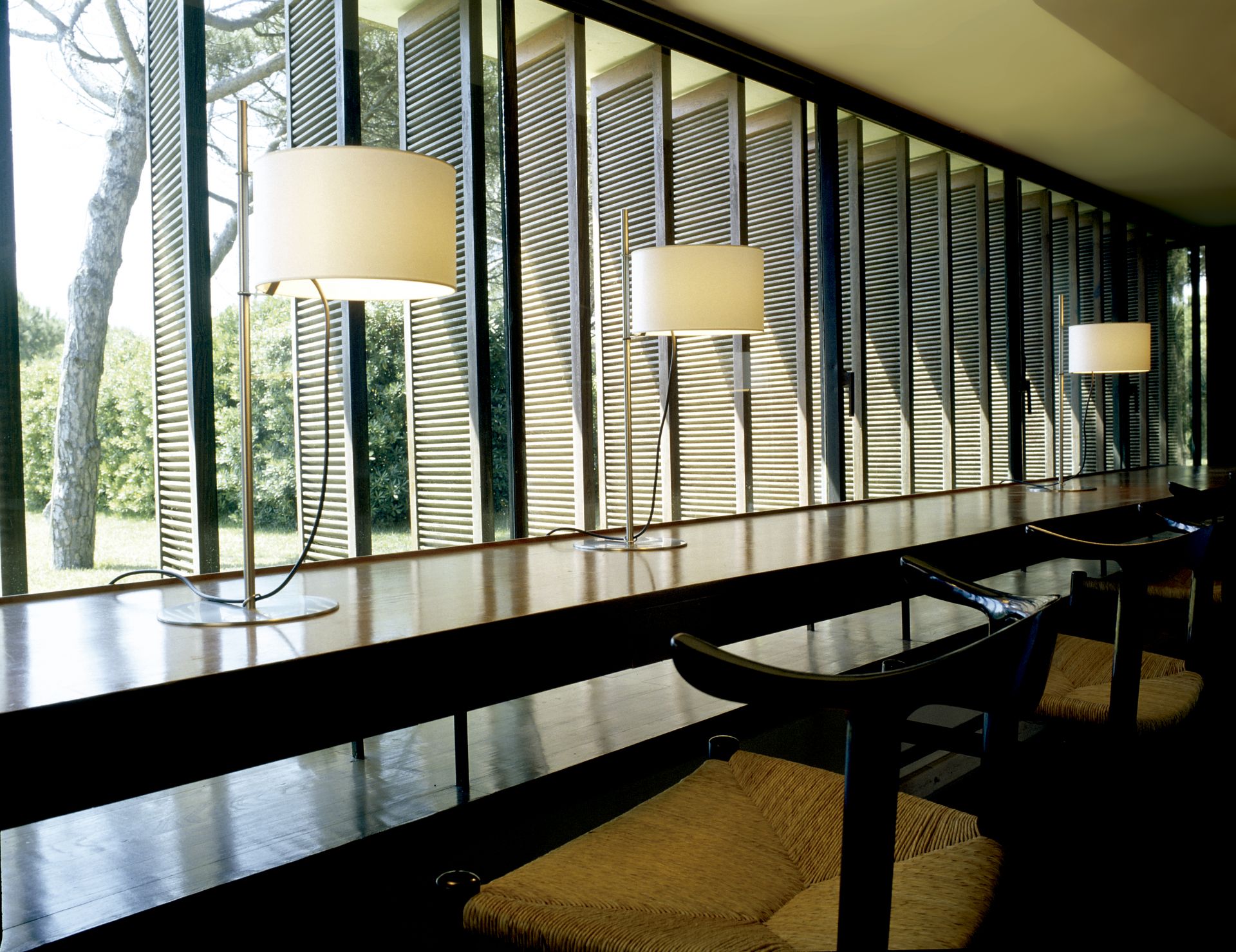 TMD Tischleuchte von Santa & Cole auf langem Holztisch vor Fenster mit Jalousien.