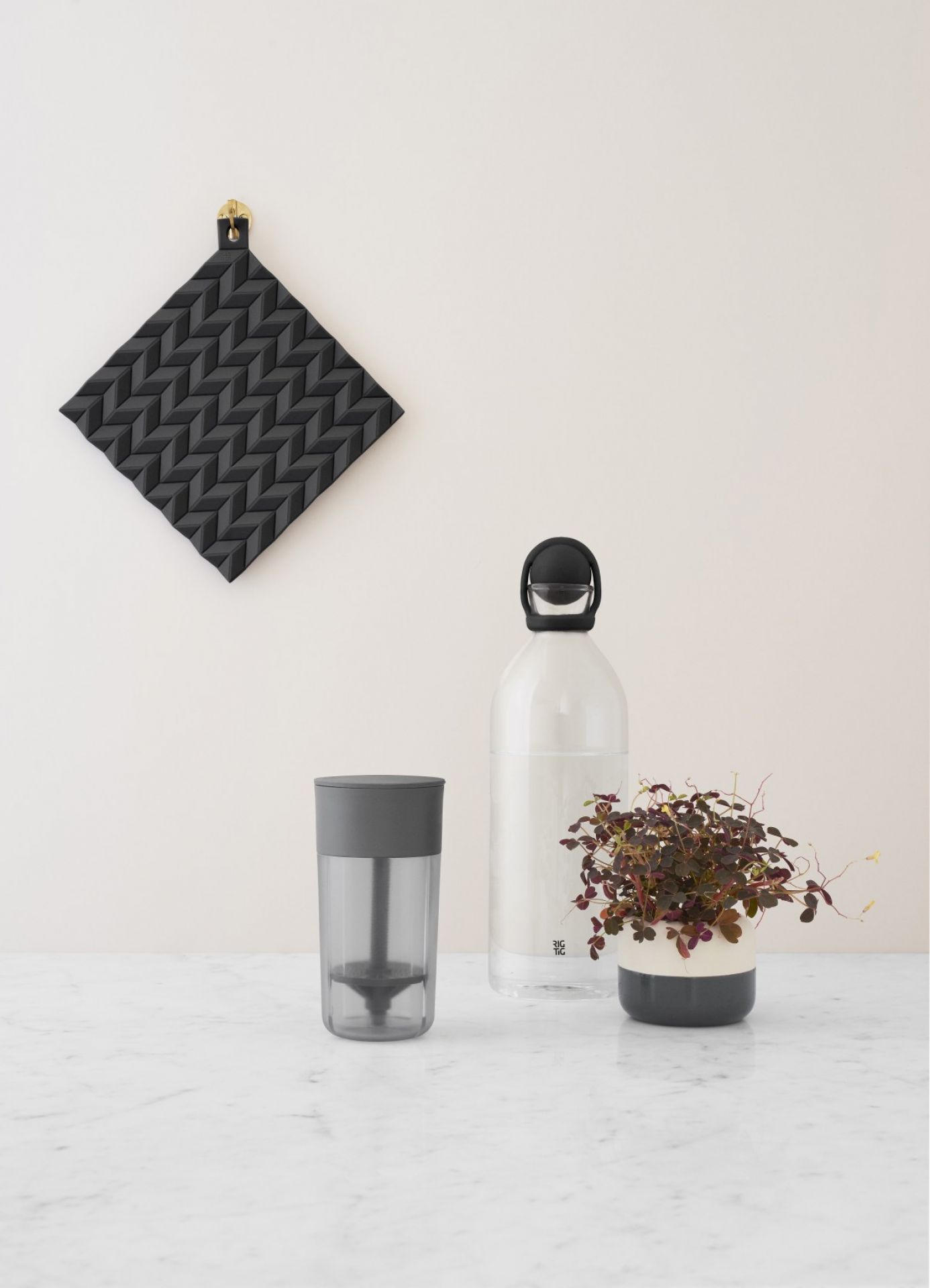 Stillleben mit RIG-TIG Wasserkaraffe, schwarzem Topflappen und Pflanze auf Marmortisch. Küchendekoration und Tischaccessoires.